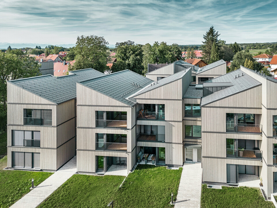 Vue aérienne de l'immeuble d'appartements moderne du quartier Schmucker à Utting am Ammersee, avec une construction hybride en bois durable et d'élégantes façades en bois. Les bâtiments sont équipés de R.16 PREFA durables en gris sombre P.10. Cette architecture innovante, conçue par WWA Architects de Munich, se caractérise par des lignes claires et une intégration harmonieuse dans l'environnement verdoyant. Les balcons spacieux et les grandes fenêtres assurent une atmosphère de vie lumineuse et conviviale, tandis que les matériaux de haute qualité garantissent durabilité et résistance aux intempéries.