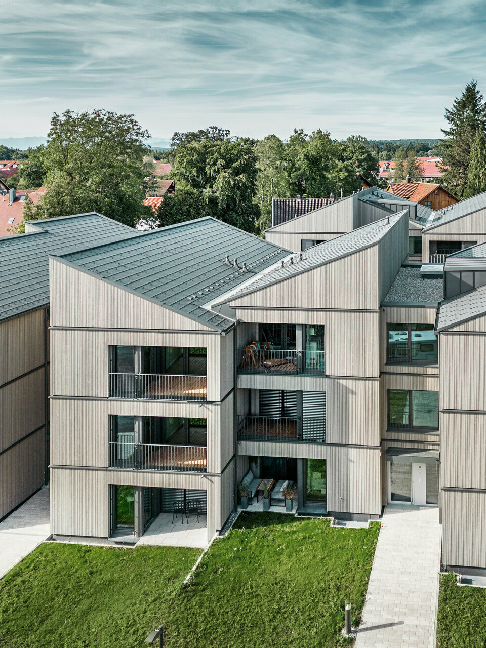 Vue aérienne de l'immeuble d'appartements moderne du quartier Schmucker à Utting am Ammersee, avec une construction hybride en bois durable et d'élégantes façades en bois. Les bâtiments sont équipés de R.16 PREFA durables en gris sombre P.10. Cette architecture innovante, conçue par WWA Architects de Munich, se caractérise par des lignes claires et une intégration harmonieuse dans l'environnement verdoyant. Les balcons spacieux et les grandes fenêtres assurent une atmosphère de vie lumineuse et conviviale, tandis que les matériaux de haute qualité garantissent durabilité et résistance aux intempéries.