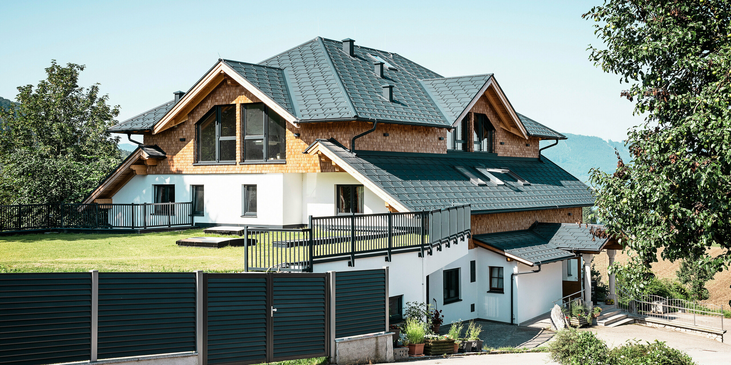 Vue latérale du café-restaurant Zenz'n Stub'n à Schörfling am Attersee avec un toit moderne en aluminium composé de panneaux de toiture PREFA R.16 avec revêtement de haute qualité P.10 anthracite. Le toit durable PREFA allie esthétique et fonctionnalité et offre une protection optimale. L'accès au jardin et l'intégration harmonieuse du bâtiment dans l'environnement naturel soulignent le design intemporel.