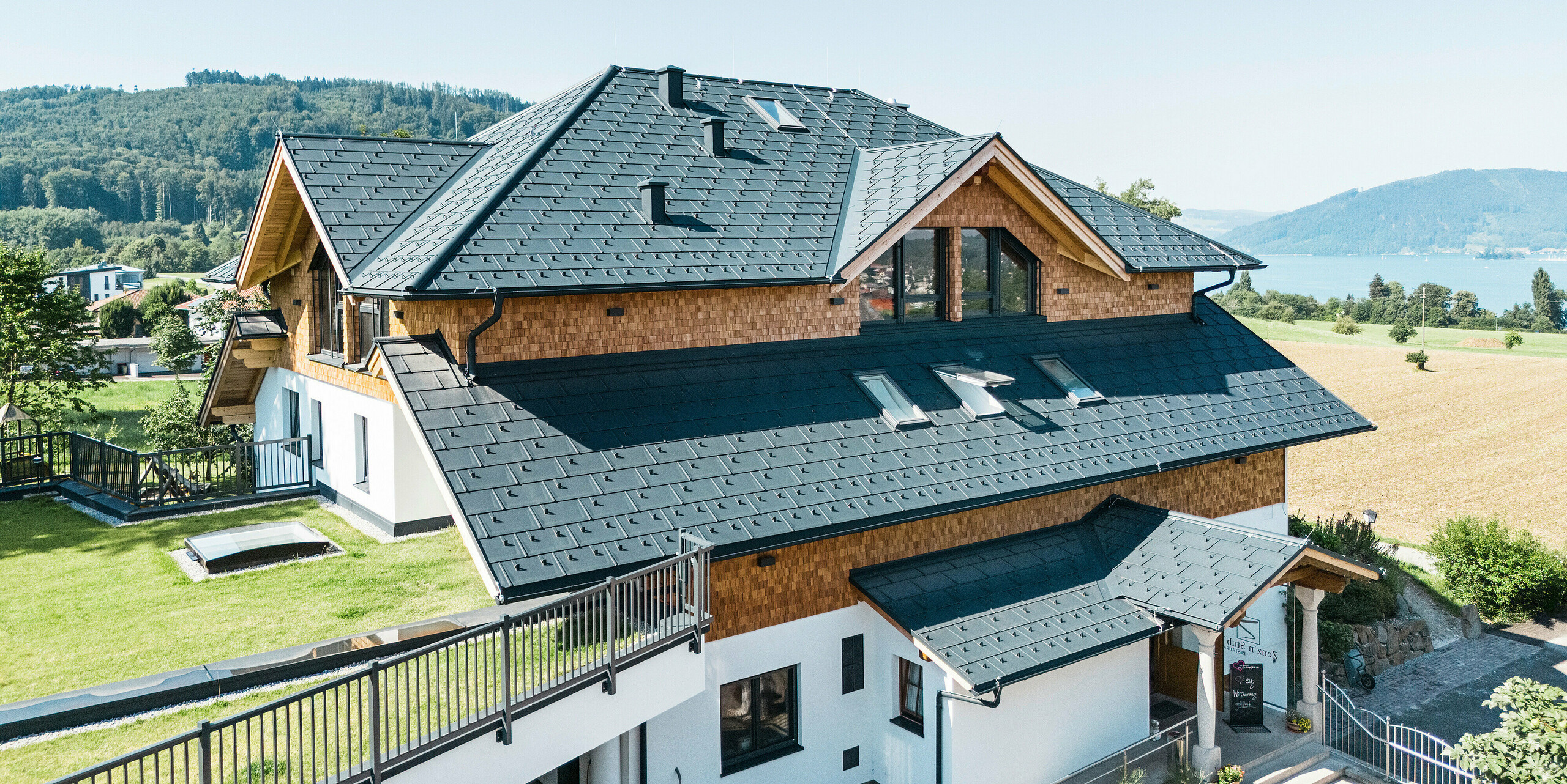 Vue aérienne du café-restaurant Zenz'n Stub'n à Schörfling am Attersee, dont la toiture a été refaite avec des panneaux de toiture PREFA R.16 en P.10 anthracite. Le toit en aluminium durable confère au bâtiment un aspect moderne et élégant tout en offrant une protection robuste. Le drainage du toit harmonieusement intégré et l'environnement naturel complètent le tableau d'ensemble.