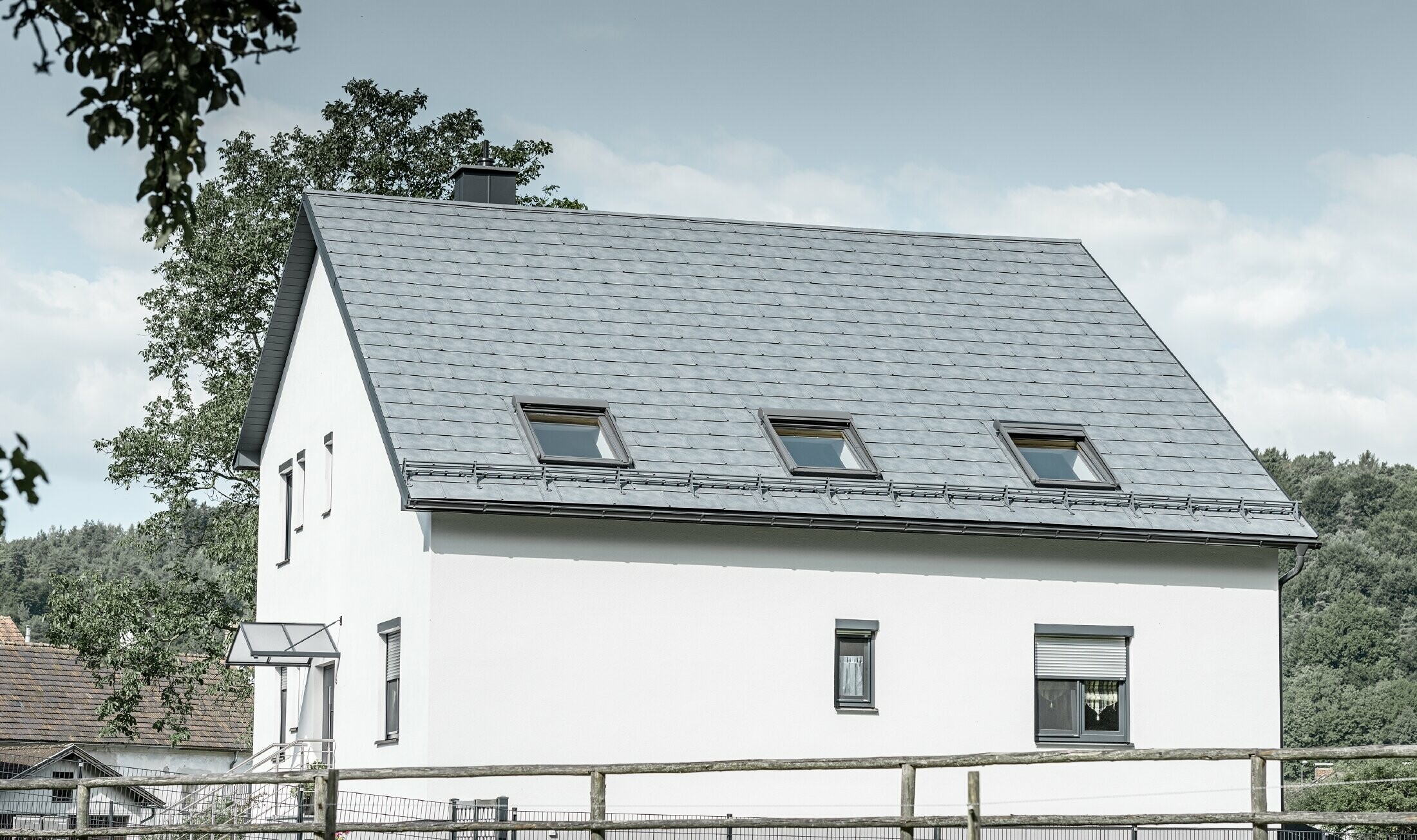 Le toit à deux pans de la maison individuelle classique a été recouvert avec la nouvelle tuile R.16 PREFA couleur gris pierre. Trois fenêtres de toit ont été intégrées dans la surface de toit et des arrêts neige ont été montés. La façade a été simplement laissée blanche.