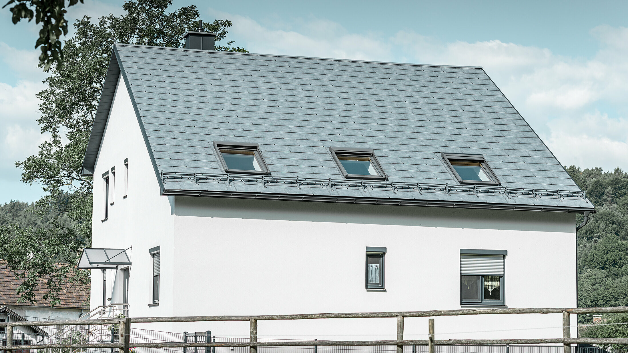 Le toit à deux pans de la maison individuelle classique a été recouvert avec la nouvelle tuile R.16 PREFA couleur gris pierre. Trois fenêtres de toit ont été intégrées dans la surface de toit et des arrêts neige ont été montés. La façade a été simplement laissée blanche.