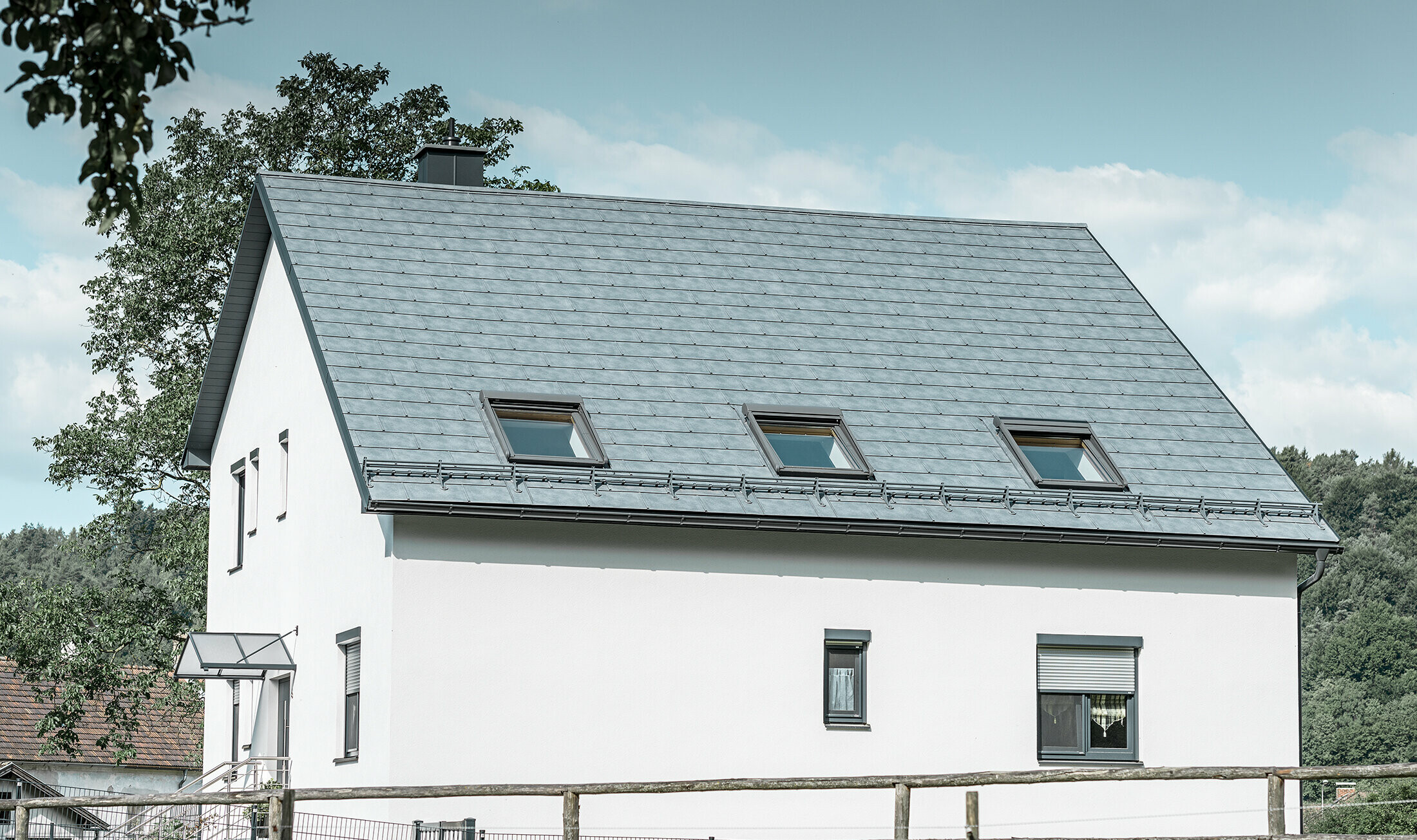 Le toit à deux pans de la maison individuelle classique a été recouvert avec la nouvelle tuile R.16 PREFA couleur gris pierre. Trois fenêtres de toit ont été intégrées dans la surface de toit et des arrêts neige ont été montés. La façade a été simplement laissée blanche.