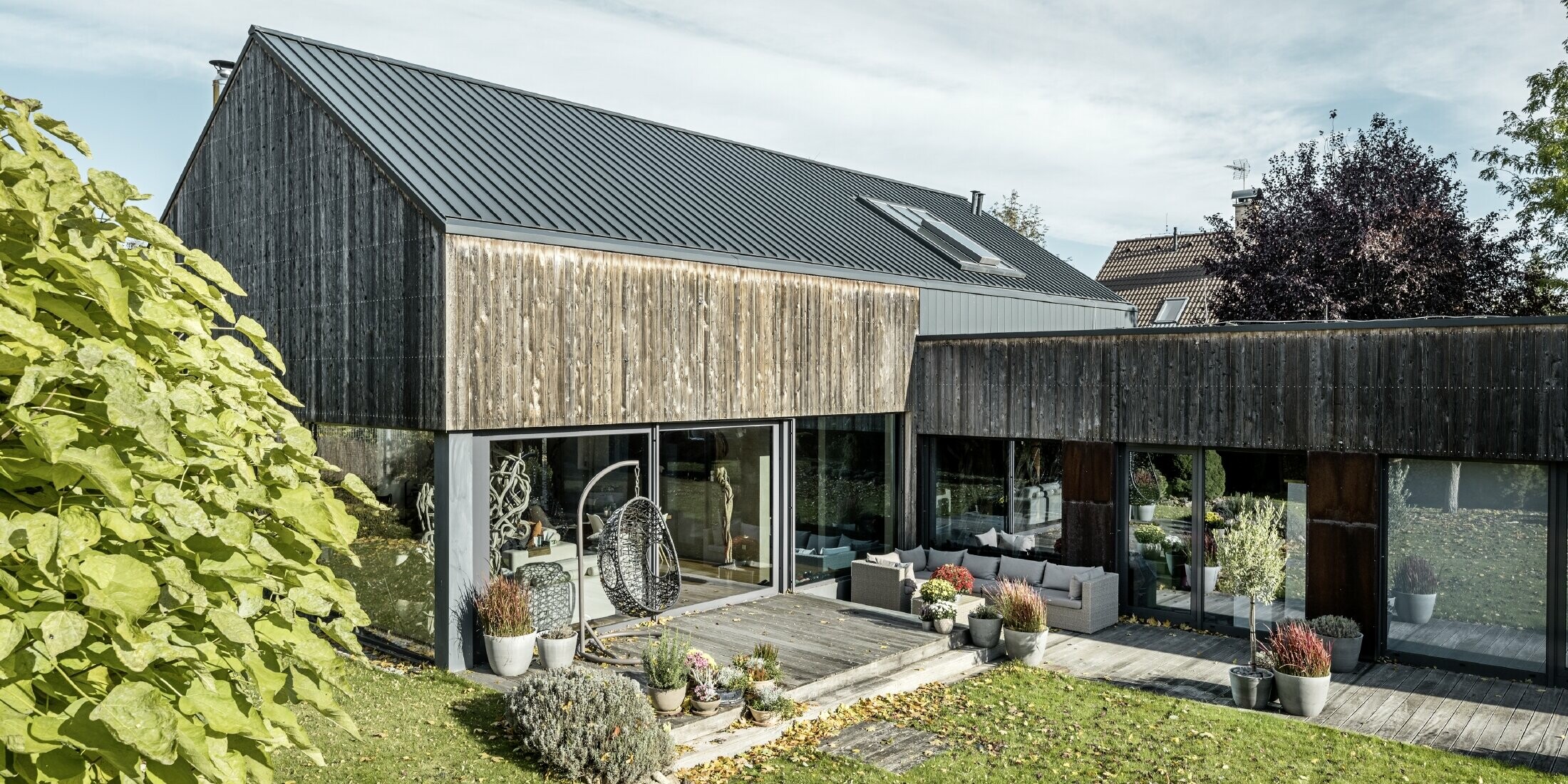 Maison individuelle avec toit à pignon, recouvert avec les joints debout double agrafe PREFALZ en anthracite et façade en bois patiné. Avec belle terrasse en bois et grandes fenêtres au rez-de-chaussée.