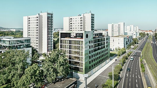 Vue aérienne de l'immeuble résidentiel et commercial "Pick-Nick" sur la Straßburger Platz à Dresde avec une façade PREFA distinctive en losanges d'aluminium dans différents tons verts et beiges. Le bâtiment est situé dans un environnement urbain moderne avec de grands immeubles et des espaces verts, à côté de la route principale. Une architecture exceptionnelle et des façades PREFA de haute qualité créent des accents visuels dans le paysage urbain.