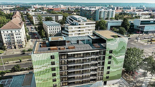 Vue aérienne de l'immeuble résidentiel et commercial "Pick-Nick" à Dresde avec une façade PREFA en losanges d'aluminium dans différents tons verts et beiges. En arrière-plan, une vue panoramique impressionnante de la ville. Le bâtiment dispose d'une spacieuse terrasse sur le toit avec des parterres de fleurs et une aire de jeux pour enfants. Situé dans un environnement urbain, il offre une combinaison d'architecture moderne, de durabilité et de design fonctionnel.