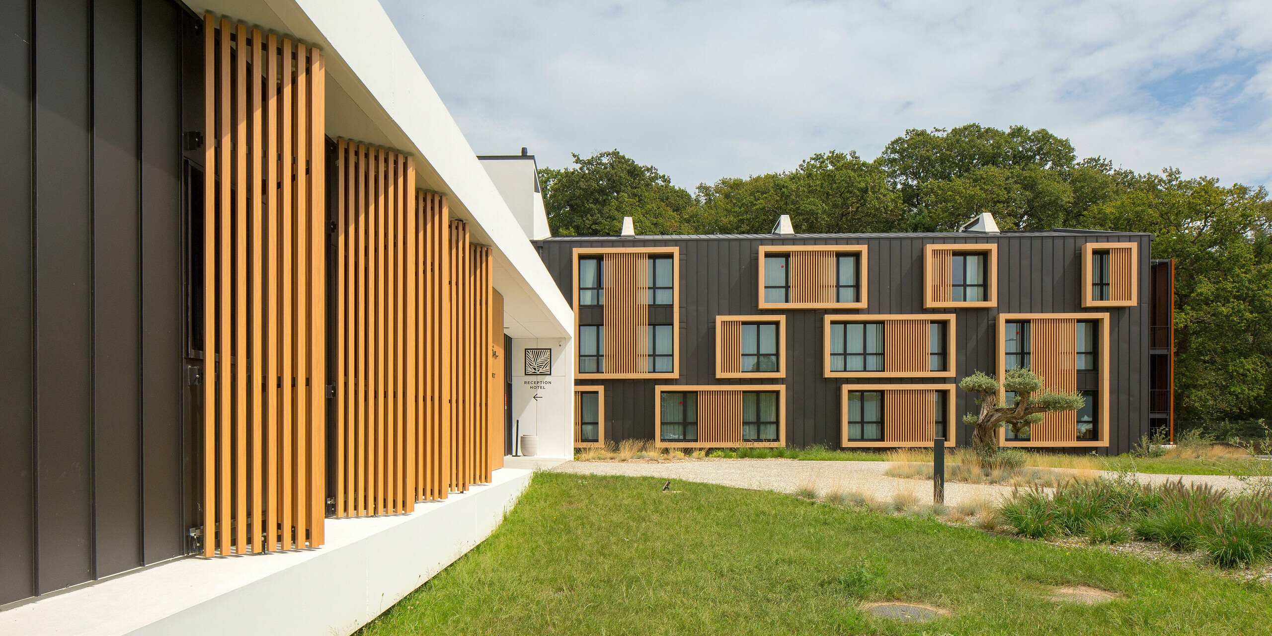 Façade moderne de l'Hôtel Parc du Landreau aux Herbiers, France, avec une combinaison de bois et d'aluminium sous la forme de PREFALZ gris noir. La conception frappante avec des éléments en bois et un toit à joint debout en tôle durable et résistante aux intempéries confère au bâtiment un caractère architectural intemporel. Des espaces verts et des fenêtres verticales soutiennent le concept de design.