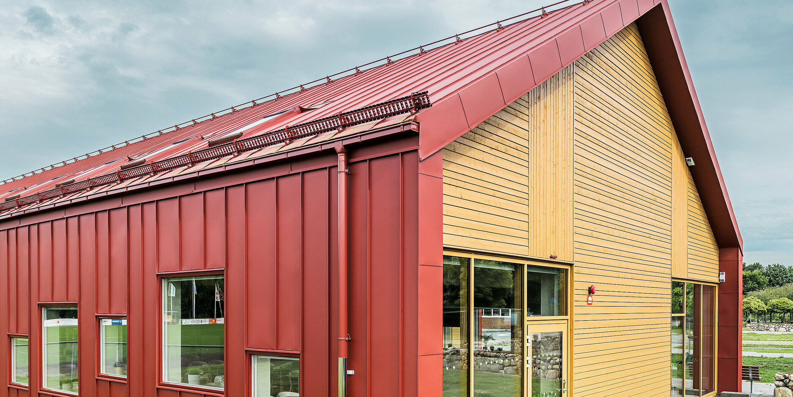 Vue latérale de la cantine scolaire Restaurang Gröna Hjärtat à Broby, Suède. La photo montre le pignon du bâtiment avec une combinaison frappante de revêtement en aluminium PREFALZ rouge oxyde et d'une façade en bois chaleureuse. Les lignes épurées et modernes de l'architecture et les contrastes de couleurs entre la toiture à joints debout rouge et le bois naturel soulignent le design innovant du bâtiment. Les fenêtres et la porte d'entrée de ce côté laissent entrer beaucoup de lumière naturelle dans les pièces et contribuent à l'atmosphère accueillante. La construction durable en tôle de haute qualité souligne la qualité et la durabilité de la structure.