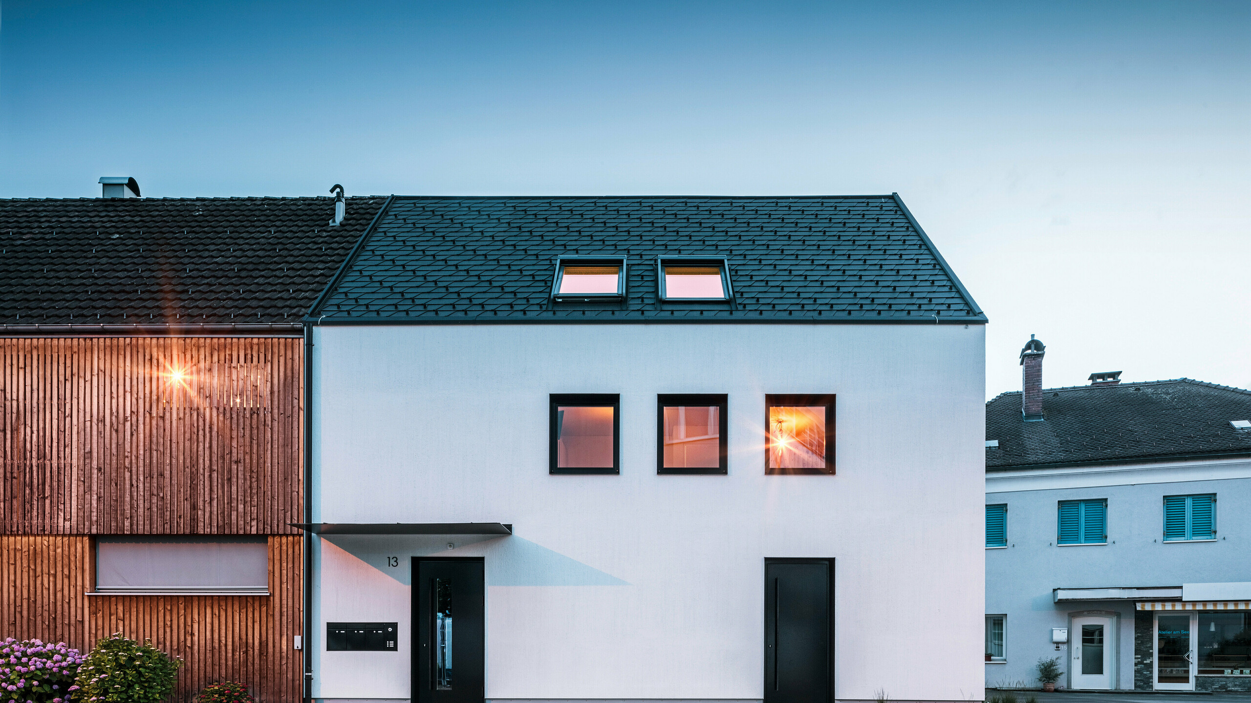 Façade moderne d'un immeuble d'habitation avec bardeaux PREFA DS19 et Prefalz en noir au bord du lac de Constance en Autriche. Le bâtiment est clairement divisé en deux, avec une extension en bois sur la gauche et une partie principale crépie sur la droite. Le toit présente la couverture en bardeaux foncés caractéristique et il y a plusieurs fenêtres rectangulaires qui se reflètent dans la chaude lumière du soir.