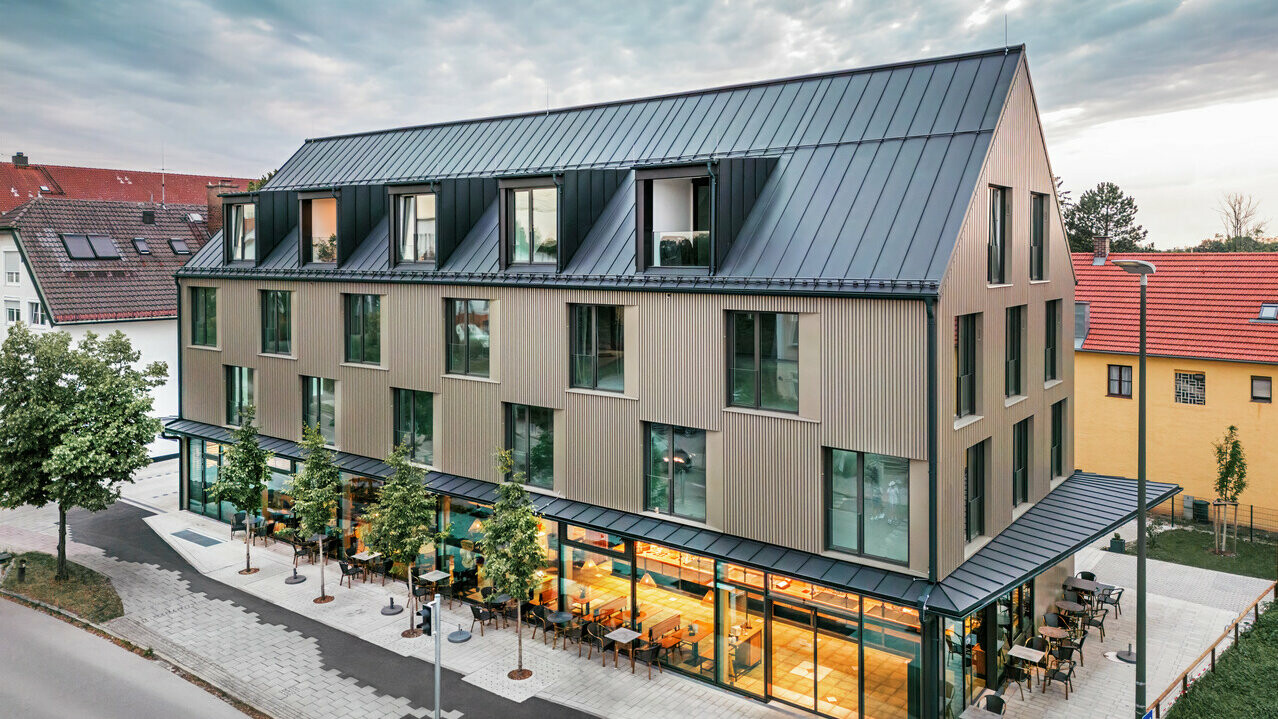 La photo montre un bâtiment moderne avec un revêtement de façade en Prefalz de couleur bronze et un toit à deux versants, également recouvert de Prefalz de couleur gris noir. La grande façade vitrée du rez-de-chaussée est encadrée d'arbres et crée une atmosphère invitante pour le restaurant situé en contrebas. La combinaison des matériaux Prefalz de haute qualité et de l'architecture moderne confère au bâtiment un aspect élégant et contemporain.