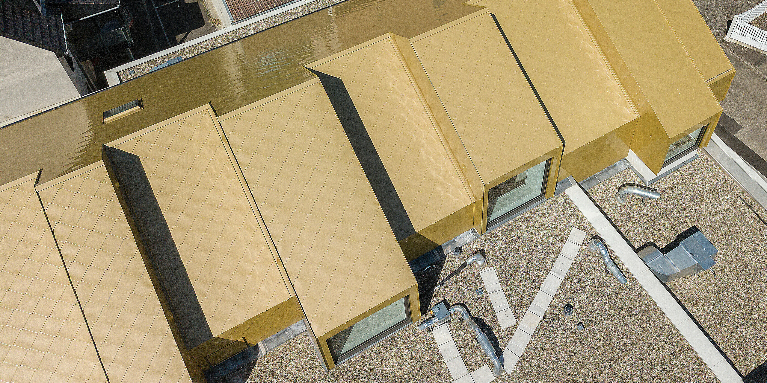 Vue d'en haut du toit doré du centre culturel de Fessenheim. Le nouveau toit a été recouvert de losanges de toiture PREFA 44 × 44 en Sparkling Gold. Les toits des bâtiments voisins sont visibles en arrière-plan.