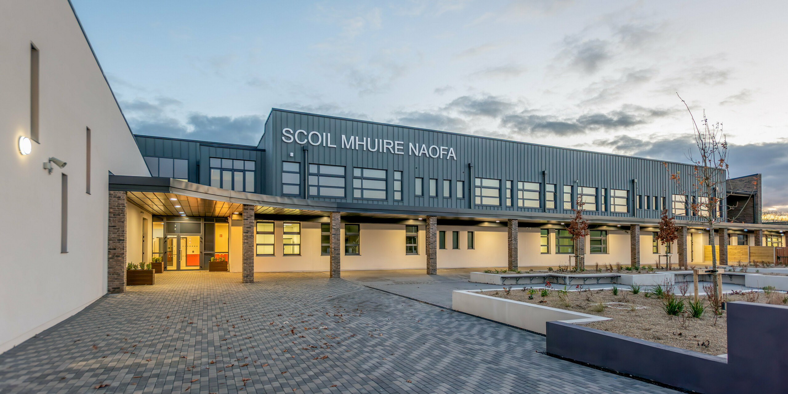 Le Scoil Mhuire Naofa à Carrigtwohill, en Irlande, au crépuscule. La façade contemporaine en aluminium PREFALZ de couleur P.10 gris souris, associée à des colonnes en briques et à des lignes claires, confère au bâtiment scolaire un aspect accueillant et de haute qualité. L'architecture claire, l'éclairage élégant et le parvis bien entretenu créent une atmosphère agréable.