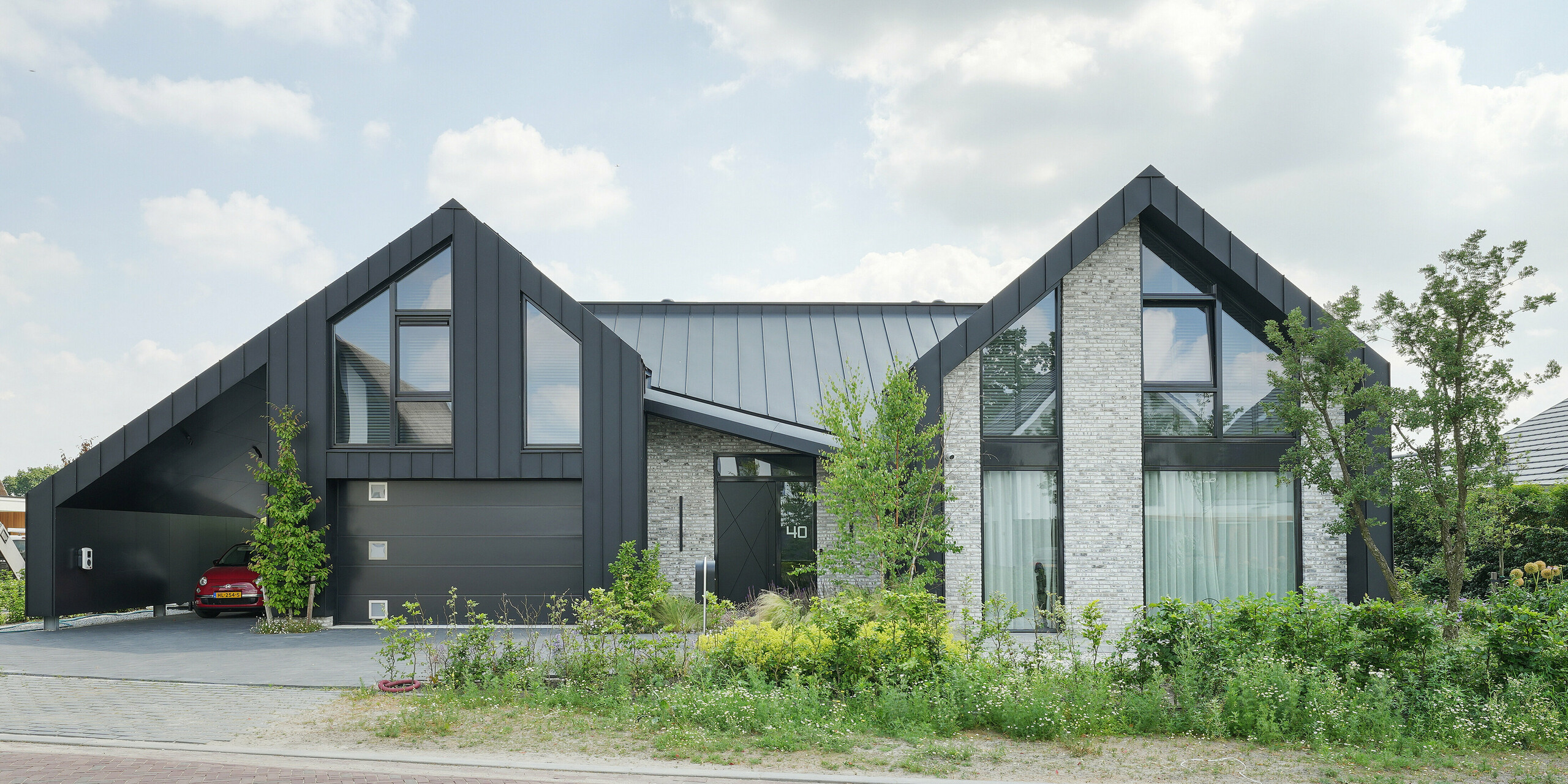 Architecture résidentielle moderne à Veenendaal, aux Pays-Bas, avec l'interaction unique de PREFALZ et d'une façade qui intègre, outre des panneaux à joint debout en aluminium, de la pierre naturelle et de grandes surfaces vitrées. La maison présente des pignons marqués et pointus qui confèrent au projet une silhouette dynamique. Le terrain est entouré d'un espace vert bien entretenu et d'une agréable voie d'accès et s'intègre harmonieusement dans un environnement calme.