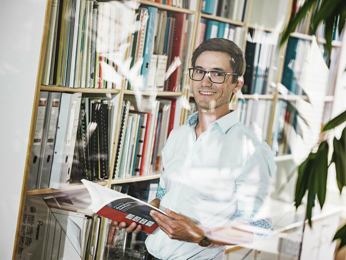 L'architecte Christof Künz, souriant dans une chemise claire, se tient dans un bureau devant une étagère remplie de classeurs et de livres. Il tient dans ses mains une brochure ouverte. La scène est animée par une plante au premier plan et par le reflet de l'intérieur du bureau sur une vitre, ce qui crée une atmosphère conviviale et riche en connaissances.