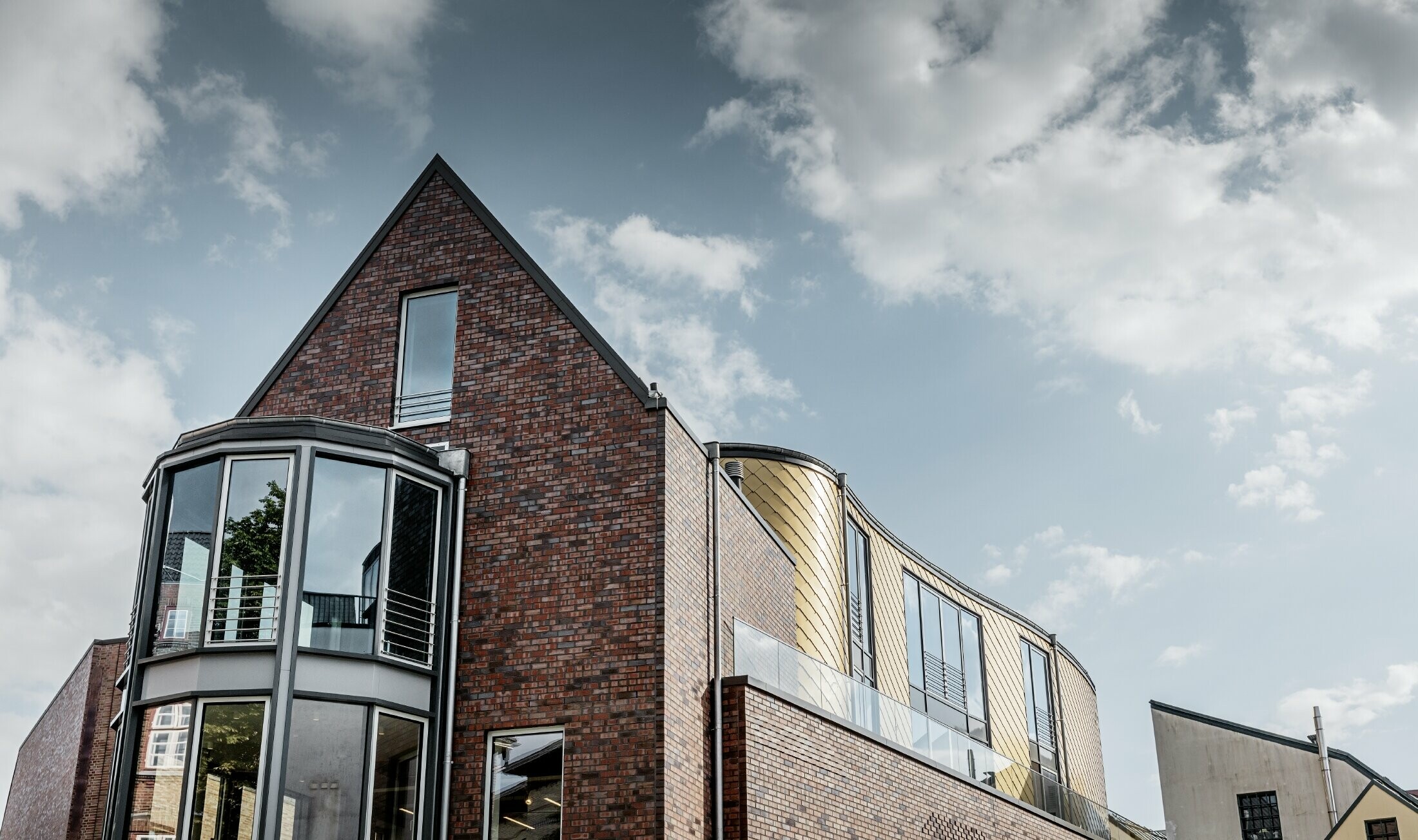 Bâtiment commercial avec façade en brique et grandes surfaces vitrées ; une partie de la façade courbe a été habillée avec le losange de façade doré en aluminium de PREFA.