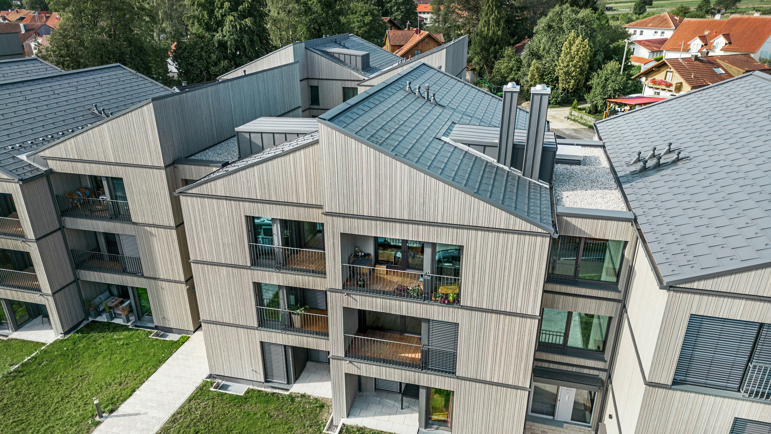 Ensemble d'appartements modernes à Utting am Ammersee, caractérisé par une construction hybride innovante avec une élégante façade en bois et des systèmes de toiture PREFA de haute qualité en aluminium. Des tuiles R.16 gris sombre et le joint debout PREFALZ ont été utilisés. Le bâtiment impressionne par ses lignes claires, ses matériaux durables et son intégration harmonieuse dans l'environnement verdoyant. La conception architecturale allie esthétique et fonctionnalité, les produits PREFA durables et résistants aux intempéries contribuant de manière significative à la haute qualité et à l'apparence impressionnante.