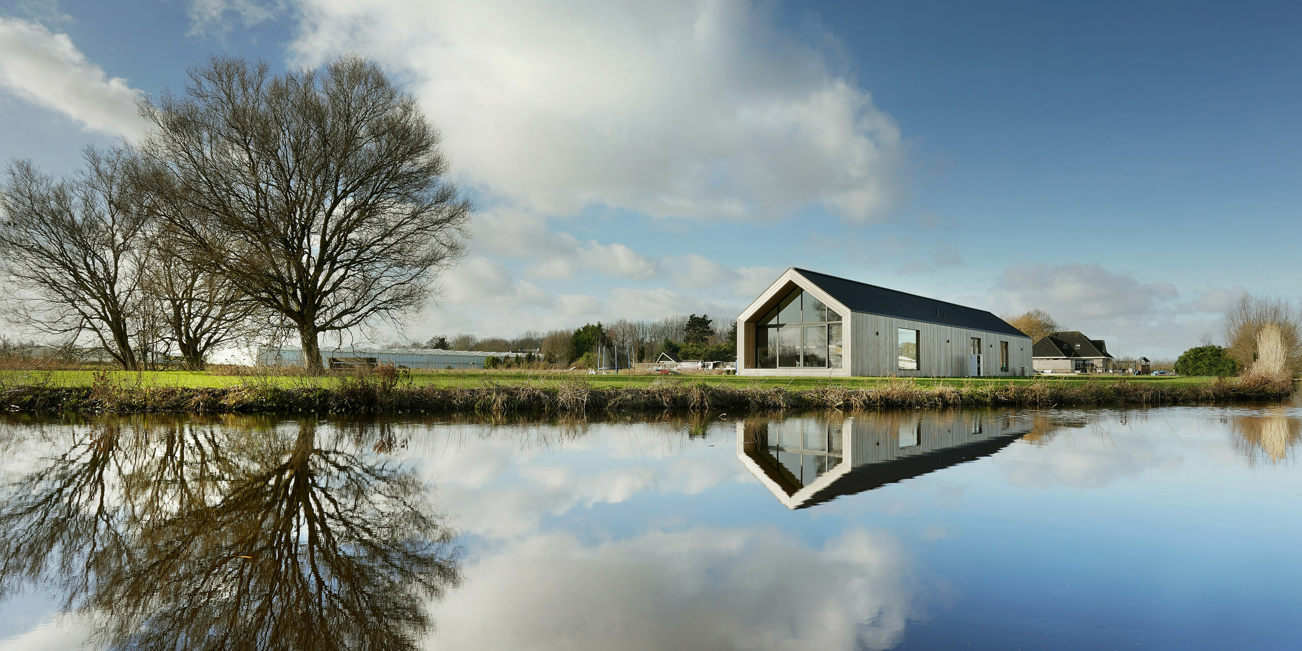 Vue panoramique d'une maison individuelle moderne au bord du lac à Uithoorn, aux Pays-Bas, avec toiture en joints debout PREFALZ en gris noir. La photo montre le bâtiment avec de grandes façades vitrées et une façade en bois naturel, qui se reflètent dans les eaux calmes du lac. Les lignes claires et les matériaux durables soulignent la construction bien pensée. Idéal pour les projets de construction innovants qui misent sur un design esthétique et une haute qualité.