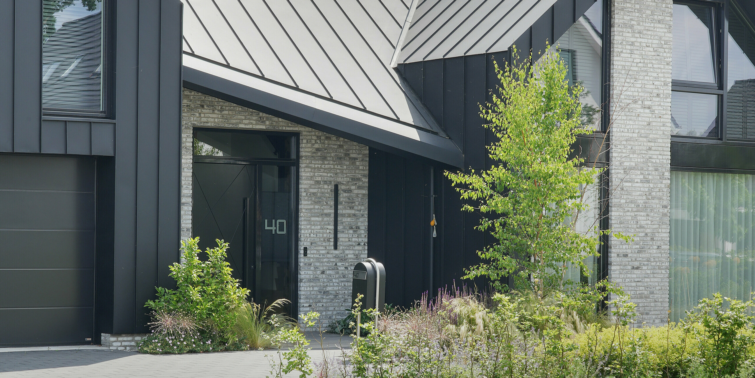 Maison individuelle moderne à Veenendaal, Pays-Bas, avec un toit noir PREFALZ. Le pignon fortement incliné et les éléments métalliques sombres se détachent remarquablement sur le ciel clair. Une pierre naturelle gris clair orne certaines parties de la façade, complétée par des fenêtres allant du sol au plafond et une porte d'entrée sobre qui confère à la maison une élégance discrète.