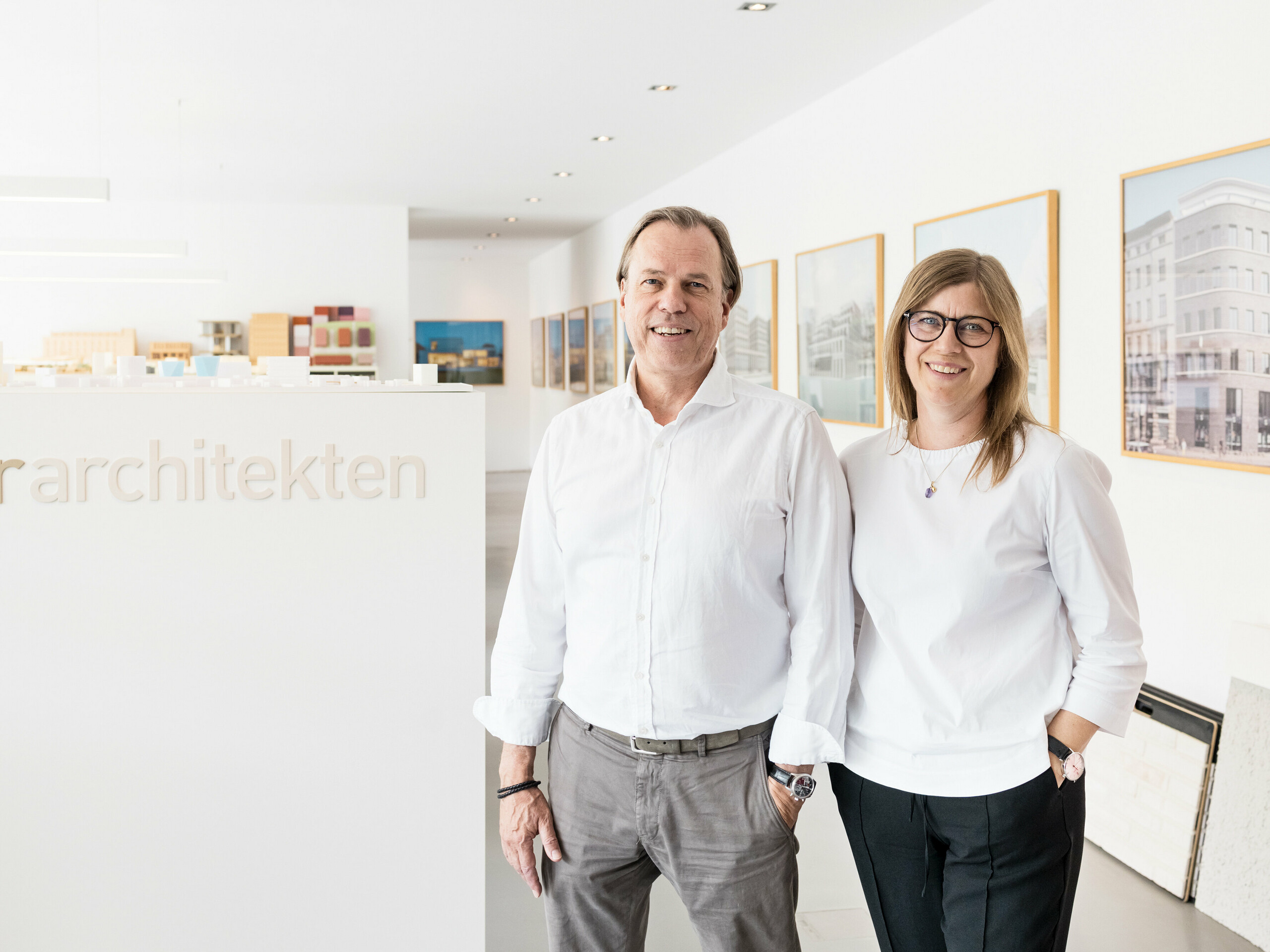 Leona et Andreas Geitner, les architectes principaux du projet à Ratingen (DE), se tiennent souriants devant un mur blanc portant l'inscription « Geitner Architects ». Tous deux portent des chemises blanches, donnant à l’image une apparence professionnelle et uniforme. En arrière-plan, vous pouvez voir des modèles architecturaux et des images encadrées de projets de construction qui reflètent l'environnement créatif et innovant du bureau d'architecture. L'atmosphère conviviale et accueillante souligne le professionnalisme et la compétence de l'équipe de Leona et Andreas Geitner.