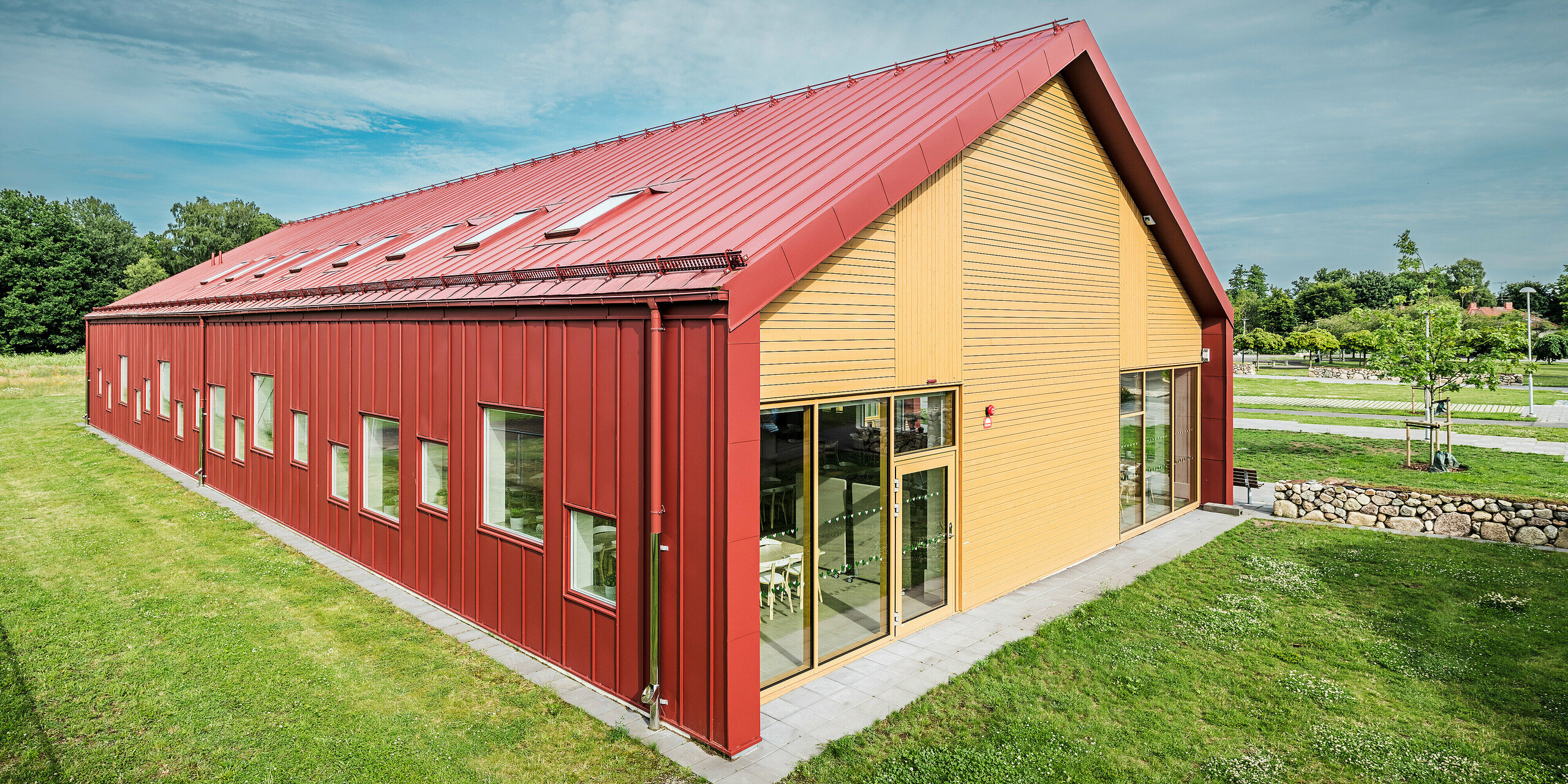 Le pignon de la cantine scolaire Gröna Hjärtat à Broby, en Suède, présente une combinaison impressionnante de PREFALZ en rouge oxyde, de bois et de généreuses surfaces en verre. Le revêtement en bois de la façade offre un contraste attrayant avec le revêtement en aluminium durable du reste du bâtiment. Les multiples fenêtres et lucarnes laissent entrer beaucoup de lumière naturelle dans le bâtiment, tandis que le design contemporain met en valeur la qualité architecturale et l'efficacité énergétique de la structure. Le bâtiment est un exemple exceptionnel d’architecture contemporaine utilisant des matériaux durables et un design innovant.