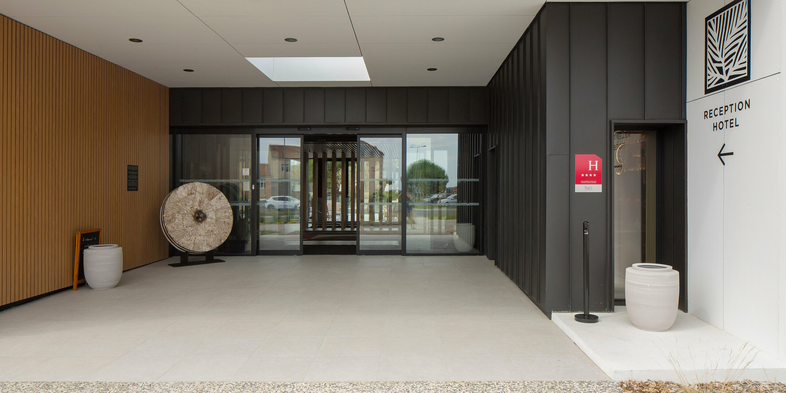 Zone d'entrée au design moderne de l'Hôtel Parc du Landreau aux Herbiers, France. La façade est revêtue de PREFALZ gris noir. Le panneau sur le bord droit de l'image indique le chemin vers la réception de l'hôtel. Des pots de fleurs, des lambris en bois et le remarquable toit en aluminium à joint debout embellissent l'entrée de l'hôtel, accessible par une porte coulissante en verre automatique.