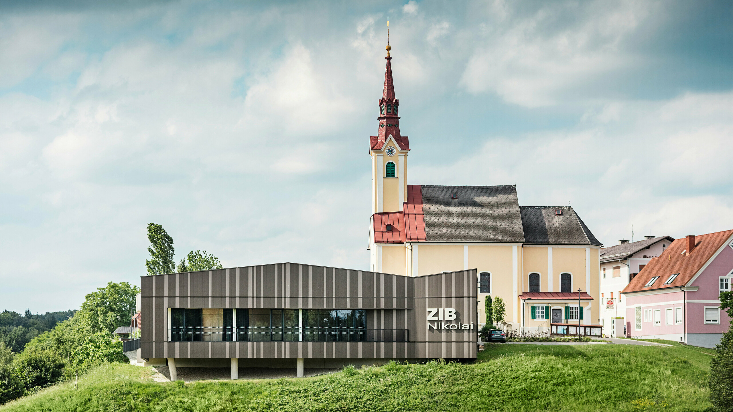 Le centre événementiel ZIB Nikolai à St. Veit dans le sud de la Styrie avec une façade en aluminium moderne et dynamique dans des tons gris brun-beige, au premier plan une prairie verte, à l'arrière-plan l'église Saint-Nicolas avec une façade jaune et un toit rouge.