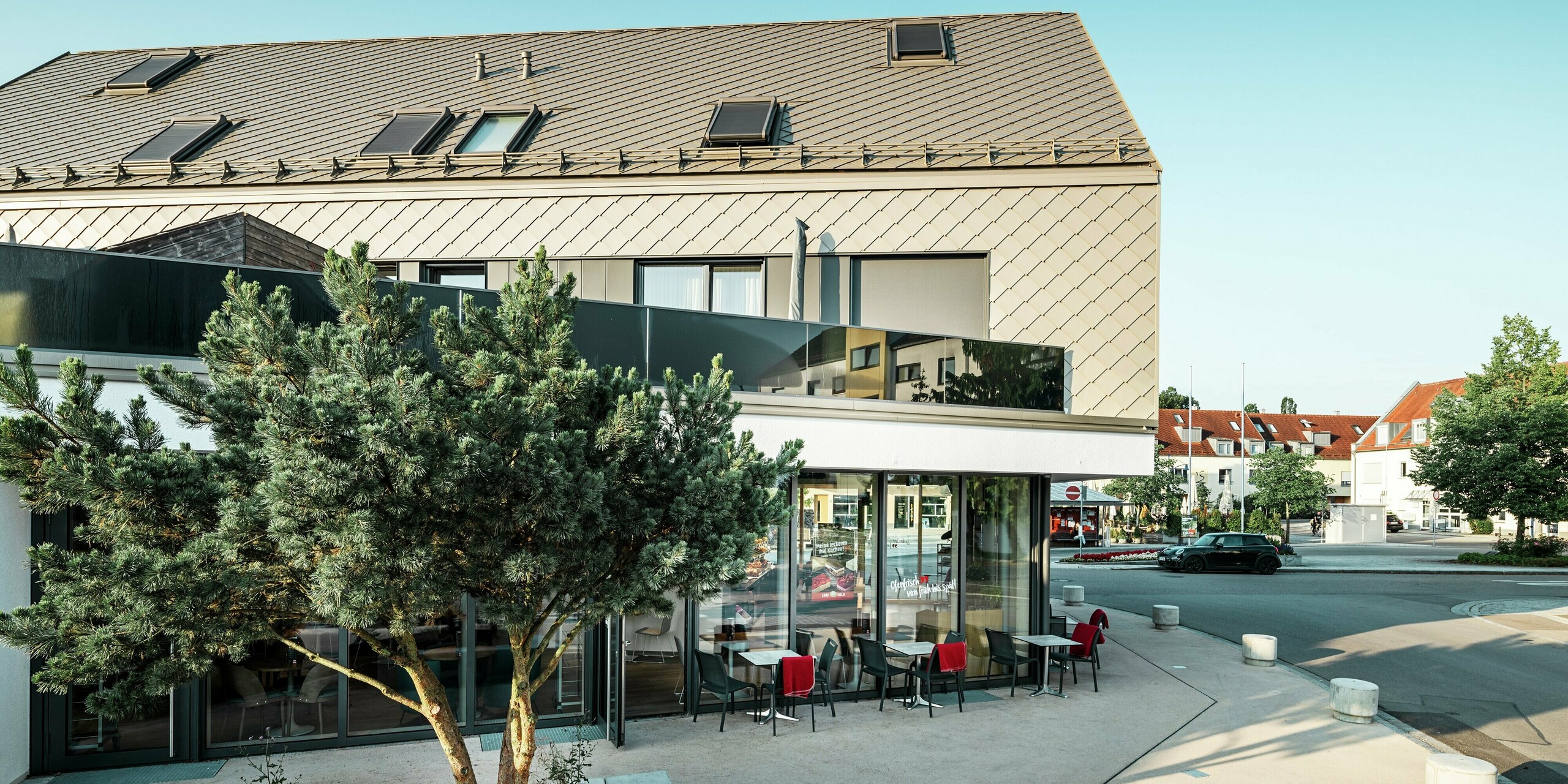 Immeuble d'appartements moderne avec une façade au design saisissant à Leitershofen, en Allemagne. Le bâtiment est recouvert de losanges de toiture et de façade 29x29 PREFA durables et de couleur bronze, qui créent un look élégant et intemporel. La combinaison de lignes claires, de grandes fenêtres et d'une façade en crépi clair au rez-de-chaussée souligne l'architecture de haute qualité. Les éléments de toiture et de façade en aluminium offrent non seulement une élégance esthétique mais également une grande résistance aux intempéries et une grande durabilité.