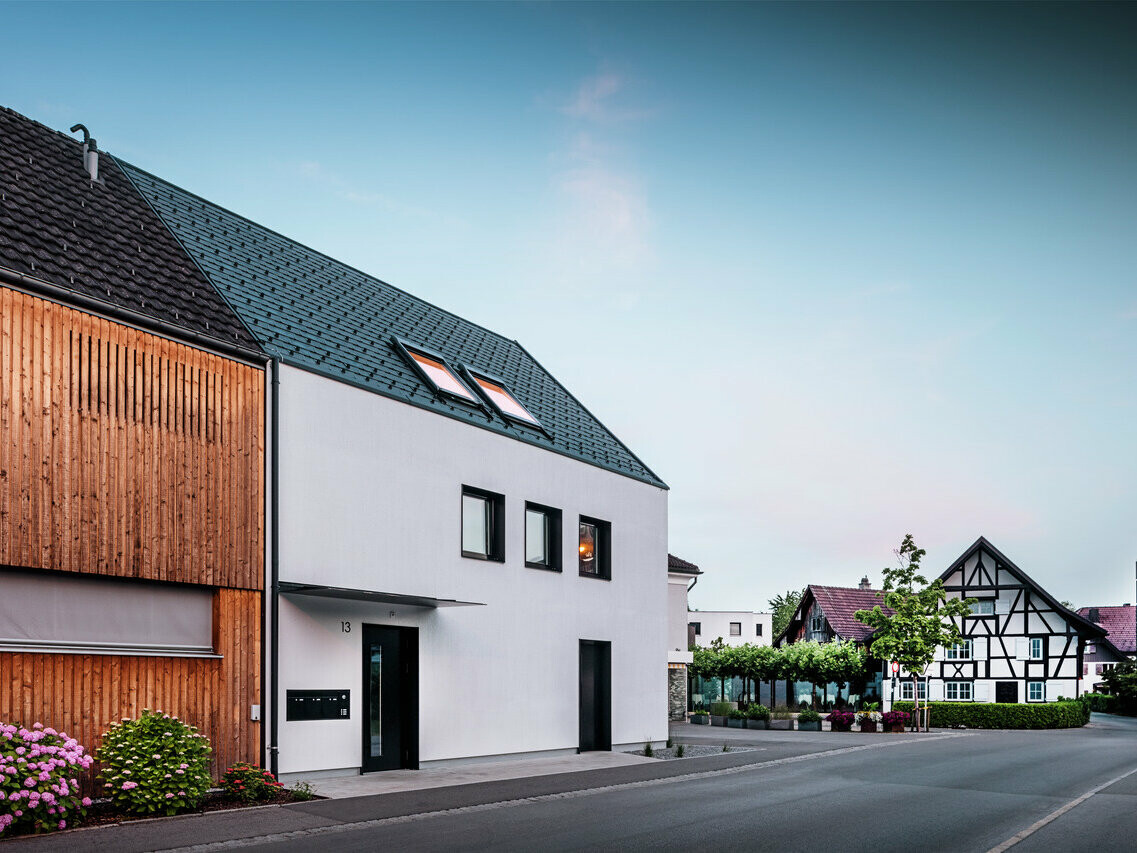 Photo d'un immeuble moderne au crépuscule, avec un toit PREFA en bardeaux noirs DS19 et prépliage. Le côté gauche de la maison est recouvert de planches de bois naturel, tandis que le côté droit est crépi et peint en blanc, avec des cadres de fenêtres noirs pour un contraste visuel fort. Au premier plan, on peut voir des arbustes en fleurs et, en arrière-plan, on distingue une maison traditionnelle à colombages, ce qui souligne le contraste harmonieux entre l'architecture moderne et traditionnelle.