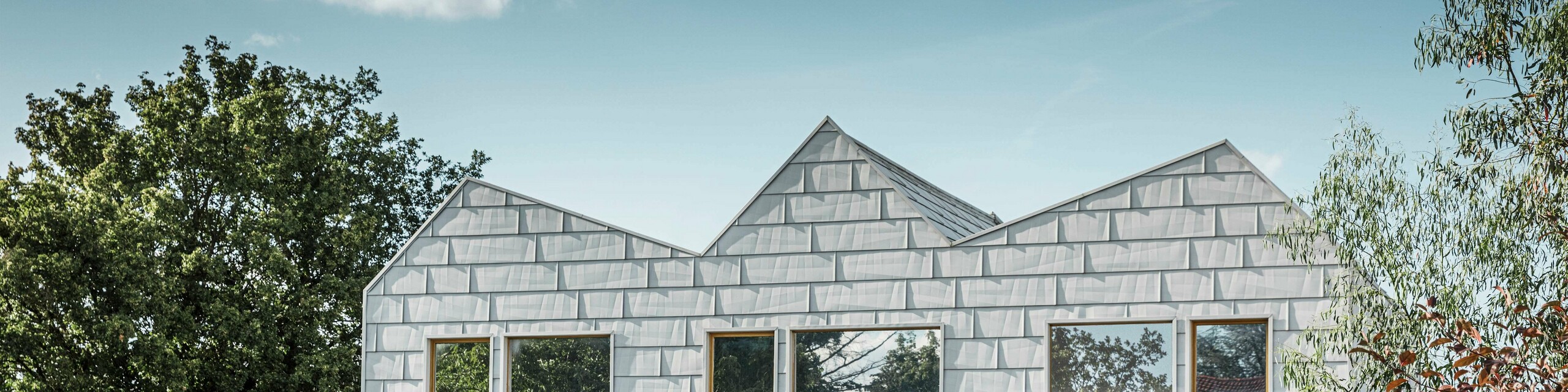 Maison jumelée moderne avec toit à pignon et façade PREFA. La façade est constituée de panneaux en aluminium FX.12, qui offrent un aspect structuré et moderne. La maison est entourée d'un jardin bien entretenu et les encadrements de fenêtres s'harmonisent parfaitement avec l'architecture globale.