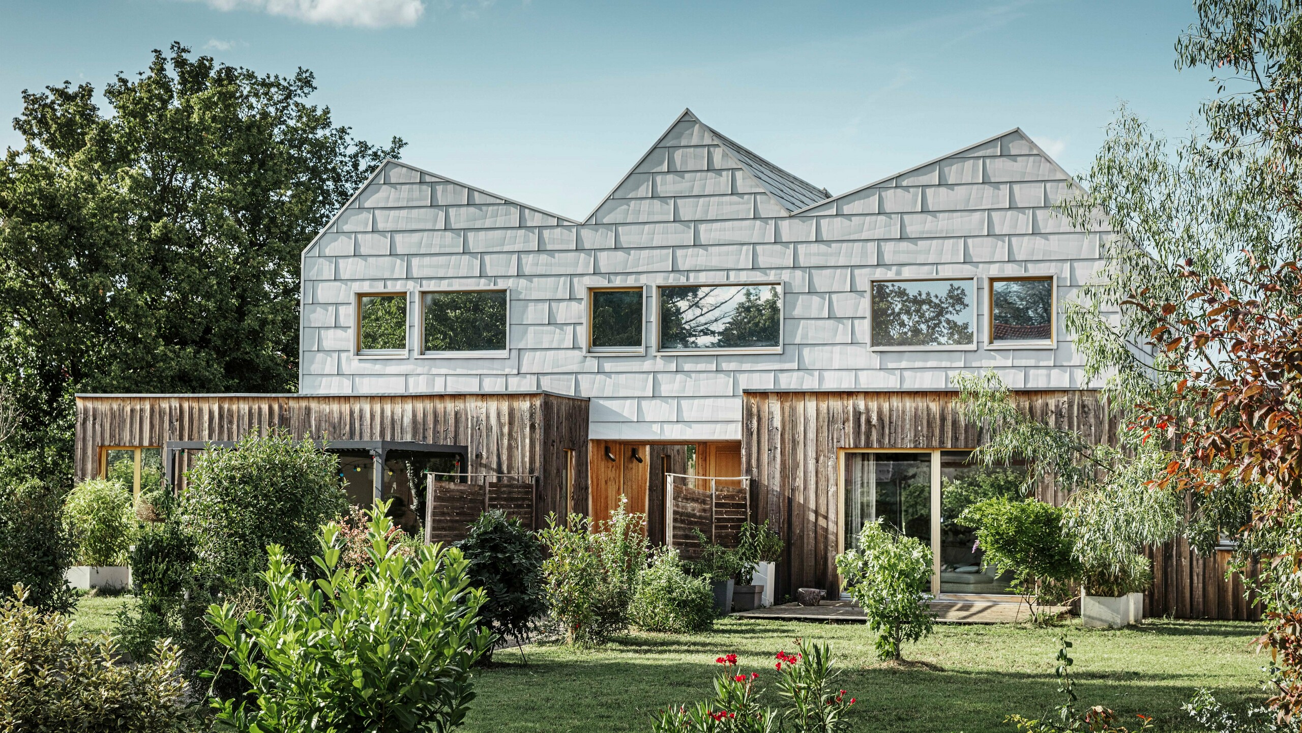 Maison jumelée moderne avec toit à pignon et façade PREFA. La façade est constituée de panneaux en aluminium FX.12, qui offrent un aspect structuré et moderne. La maison est entourée d'un jardin bien entretenu et les encadrements de fenêtres s'harmonisent parfaitement avec l'architecture globale.