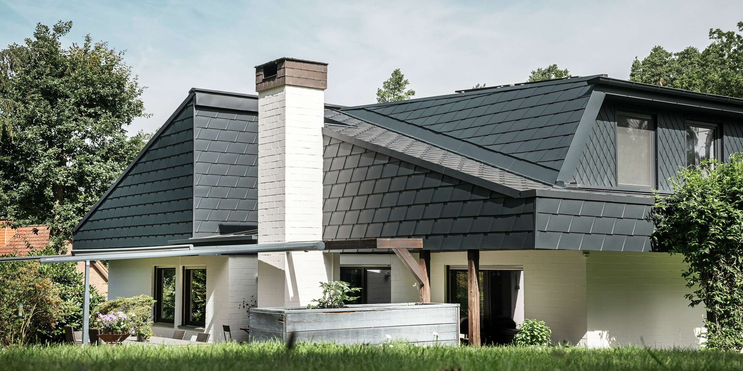 Maison individuelle à Mechernich, en Allemagne, avec DS.19 PREFA et petits losanges de toiture P.10 anthracite. La toiture en aluminium garantit une surface de toit résistante aux intempéries et durable qui s'intègre harmonieusement dans l'architecture moderne. La photo montre la maison unifamiliale traditionnelle vue de côté. Quelques arbres et une partie de la terrasse sont visibles en bord de photo.