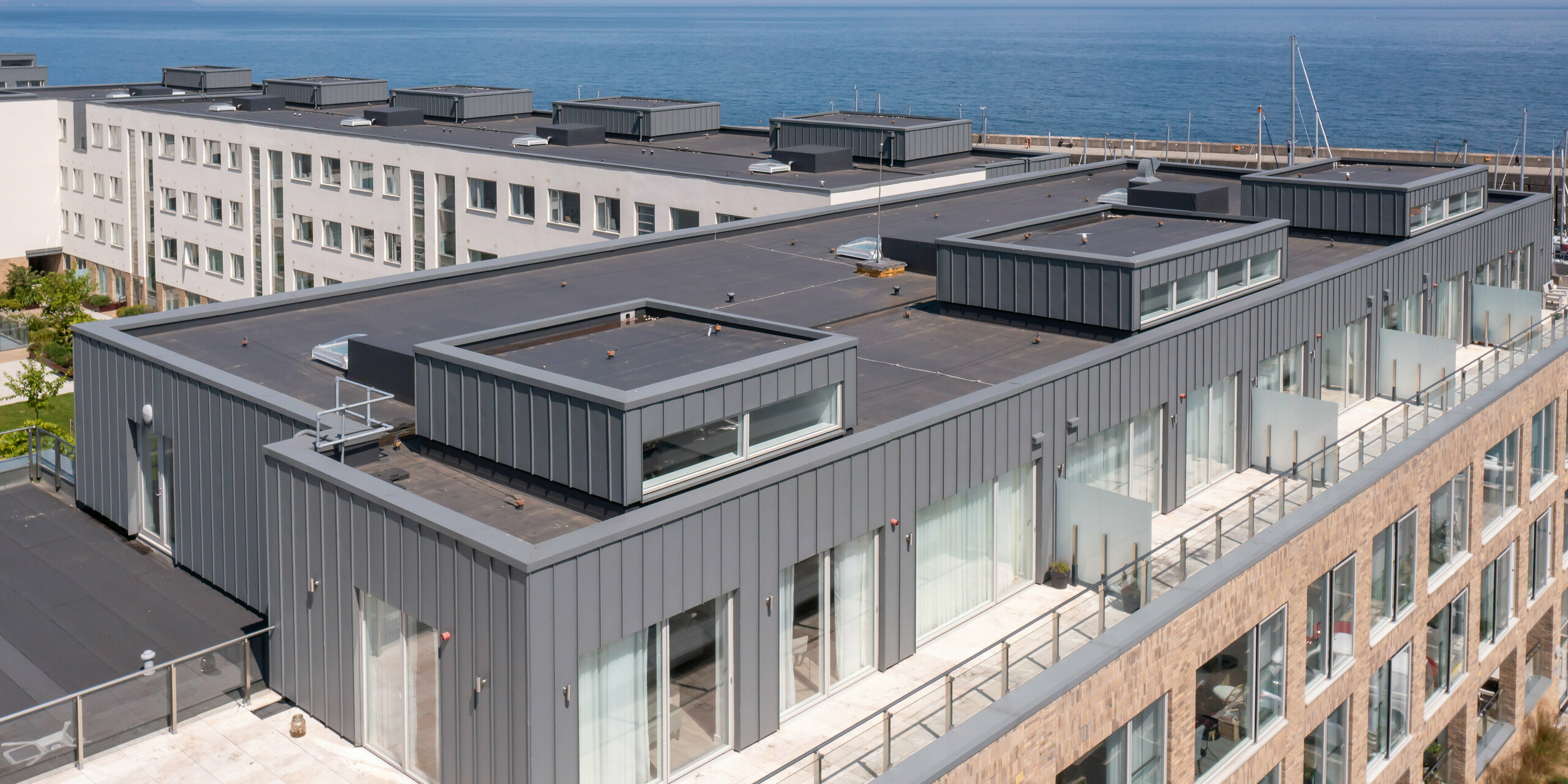 Marina Village Greystones à Wicklow, Irlande - Vue panoramique du dernier étage de l'un des bâtiments résidentiels modernes. La photo montre la vue détaillée des toits-terrasses inhabités. L'eau bleu vif de la marina est visible en arrière-plan. Le revêtement est en PREFALZ de haute qualité dans la couleur gris souris P.10. Le revêtement robuste en aluminium offre une protection durable et un aspect esthétique. De grandes baies vitrées offrent aux futurs résidents une vue imprenable sur la mer et le port de plaisance. La perspective montre délibérément l'architecture impressionnante dans le cadre pittoresque du port de plaisance.