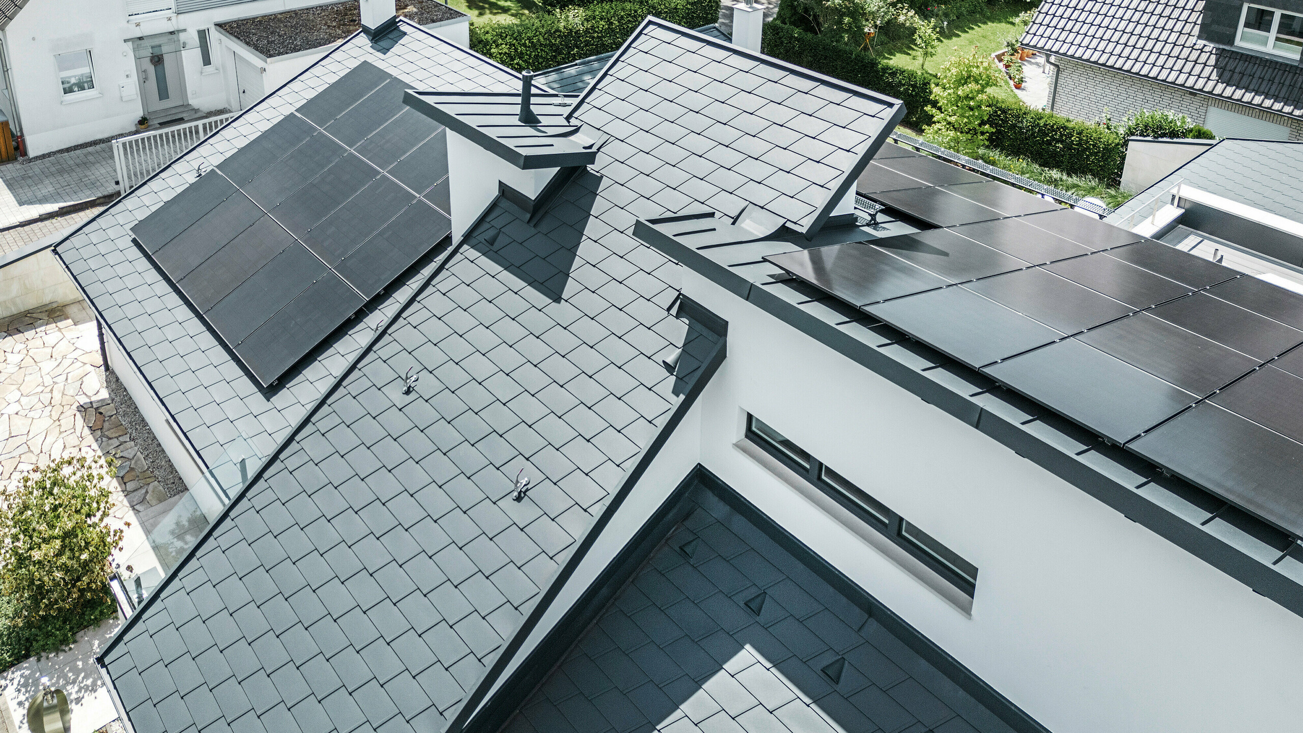 Maison individuelle moderne à Dortmund avec une installation photovoltaïque avancée, installée sur un système de toiture PREFA composé de bardeaux DS.19 et d'une couverture à joint debout PREFALZ de couleur P.10 anthracite. L'installation PV est fixée au moyen d'un support solaire Vario pour les bardeaux et d'un support solaire Vario PREFALZ pour les couvertures à joint debout. L'installation solaire garantit une production d'énergie efficace à partir de sources d'énergie renouvelables. Le design bien pensé du toit offre un équilibre optimal entre l'esthétique et la fonctionnalité.