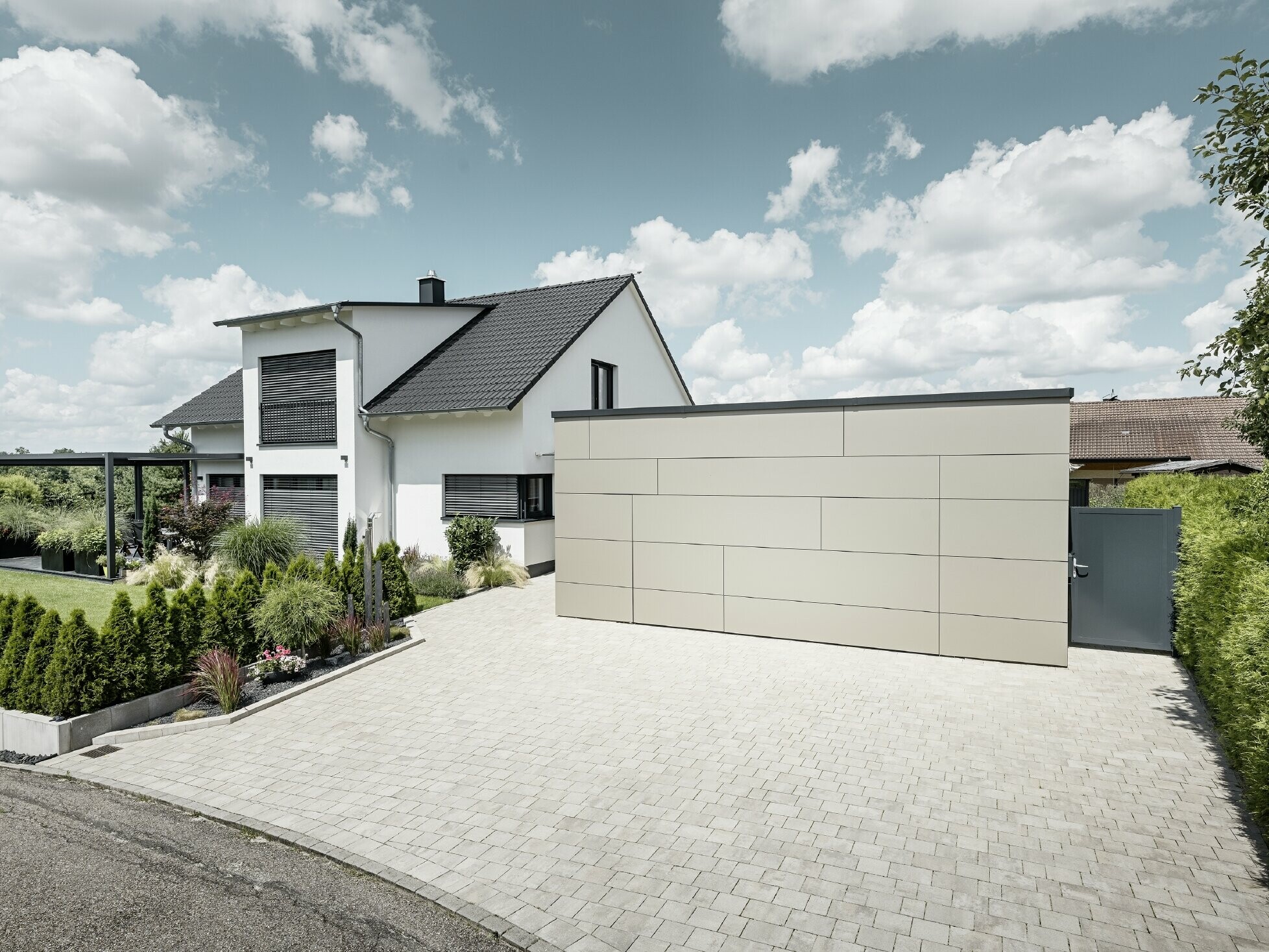 La façade du garage de cette maison individuelle est entièrement revêtue avec le panneau composite en aluminium couleur bronze. On ne distingue pas non plus la porte de garage des joints. Une allée spacieuse s'étend juste devant.