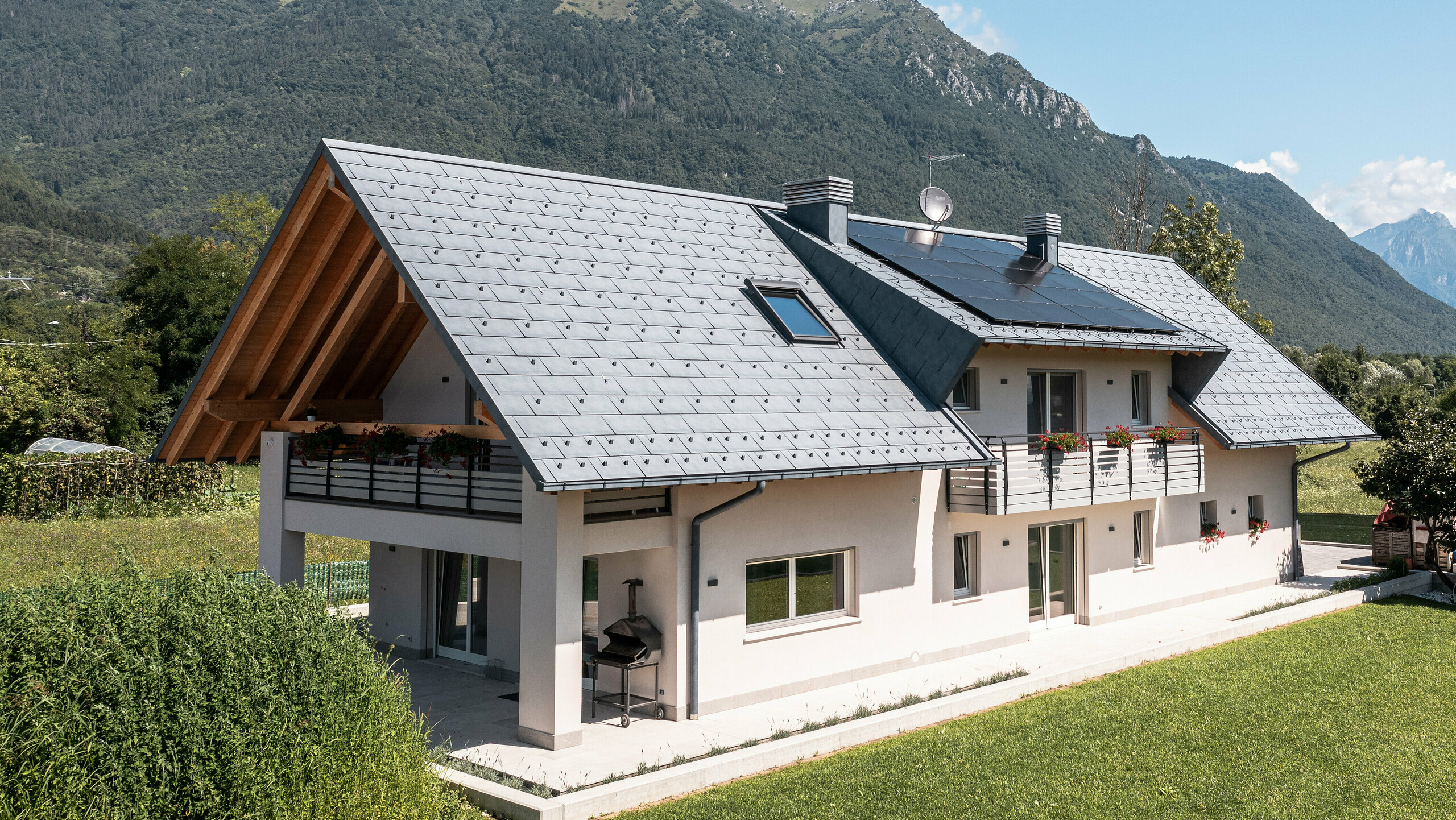 Drohnenaufnahme eines Hauses in Belluno, Italien, eingedeckt mit PREFA R.16 Dachplatten in P.10 anthrazit. Das Anwesen liegt in einer atemberaubenden Alpenlandschaft und kombiniert traditionelle Elemente wie freiliegende Holzgiebel mit modernen, langlebigen Baumaterialien, wodurch eine perfekte Symbiose aus Tradition und Moderne entsteht.