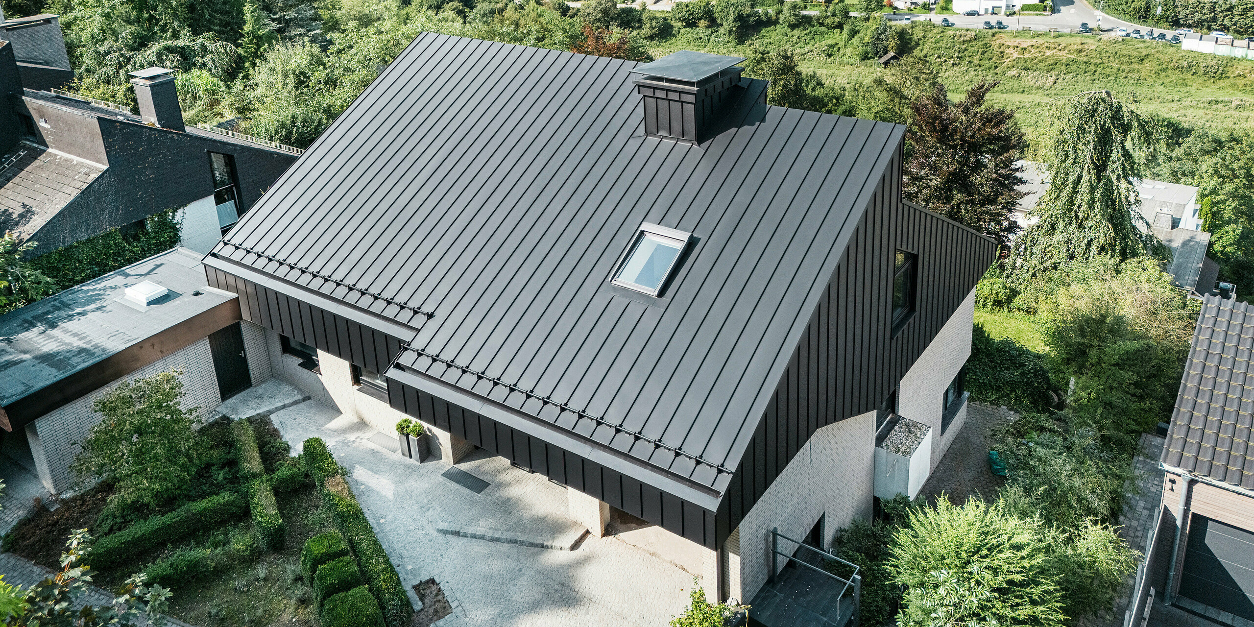 Vue de dessus d'une maison unifamiliale moderne à Meschede, en Allemagne, avec un toit en pente décalé en aluminium PREFALZ en P.10 noir. Le toit en aluminium et l'élégante façade en aluminium sur les pignons sont fabriqués avec précision et soulignent ainsi le design esthétique et résistant aux intempéries. En arrière-plan, il y a un cadre rural idyllique avec des collines verdoyantes et un quartier résidentiel calme.