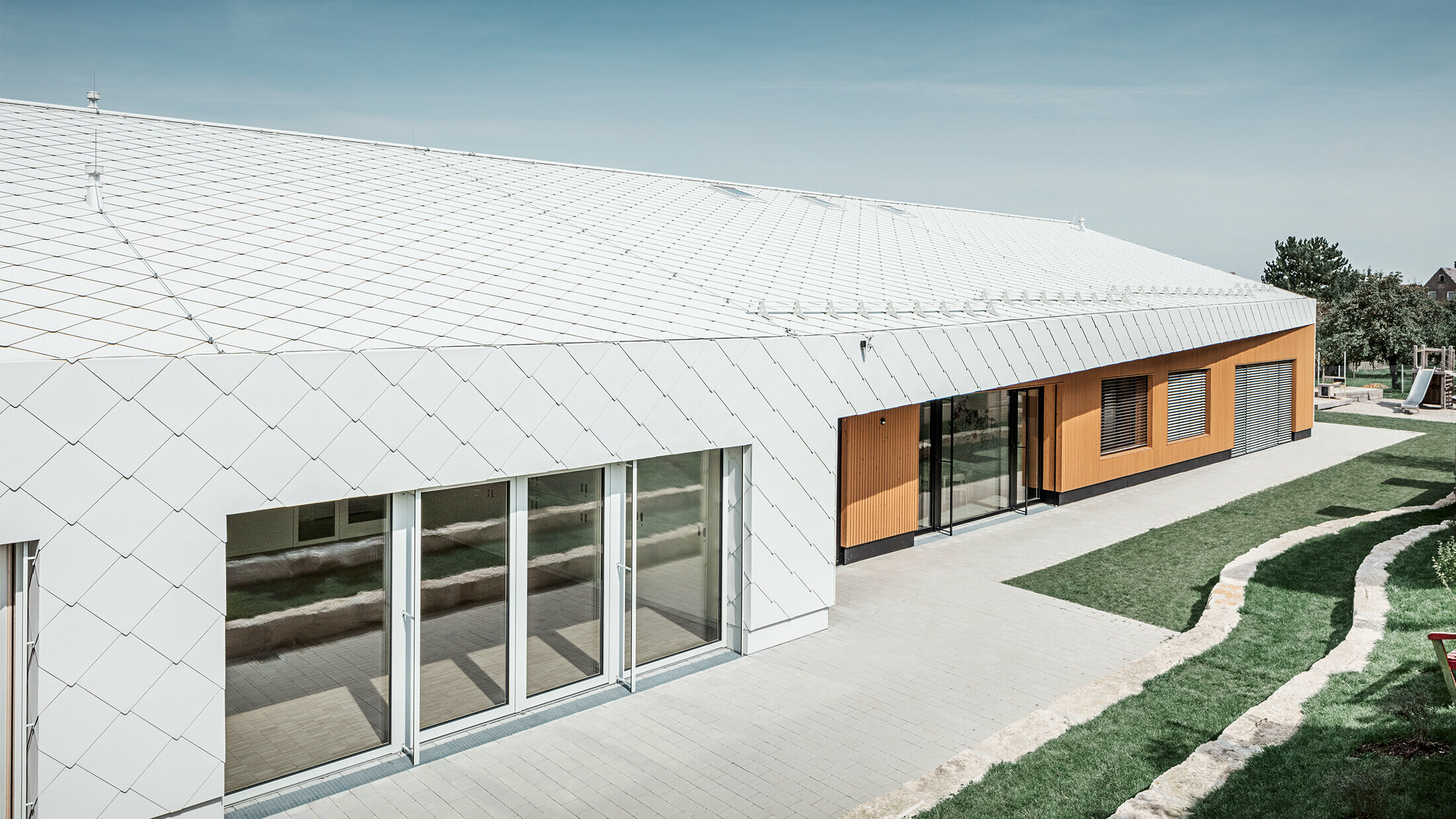 Jardin d’enfants revêtu du losange de toiture et de façade 44 de PREFA, en blanc Prefa ; la surface du toit et la façade ne font qu'un ; grandes surfaces vitrées