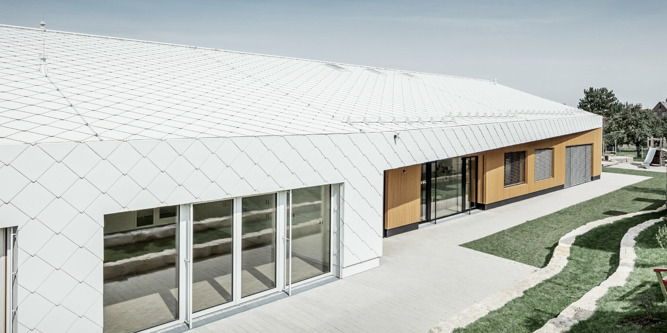 Jardin d’enfants revêtu du losange de toiture et de façade 44 de PREFA, en blanc Prefa ; la surface du toit et la façade ne font qu'un ; grandes surfaces vitrées