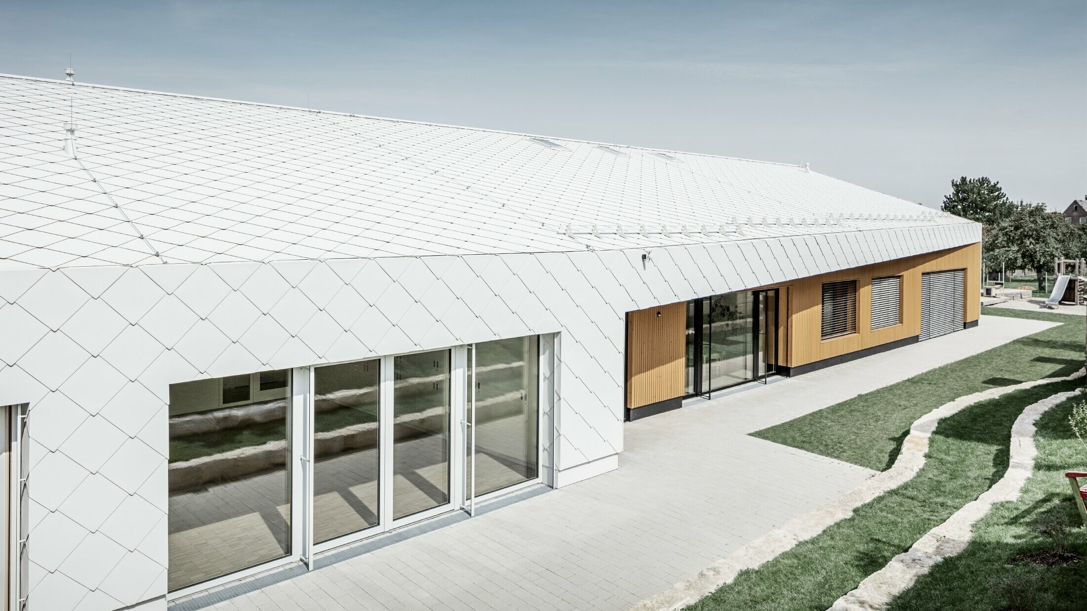 Jardin d’enfants revêtu du losange de toiture et de façade 44 de PREFA, en blanc Prefa ; la surface du toit et la façade ne font qu'un ; grandes surfaces vitrées