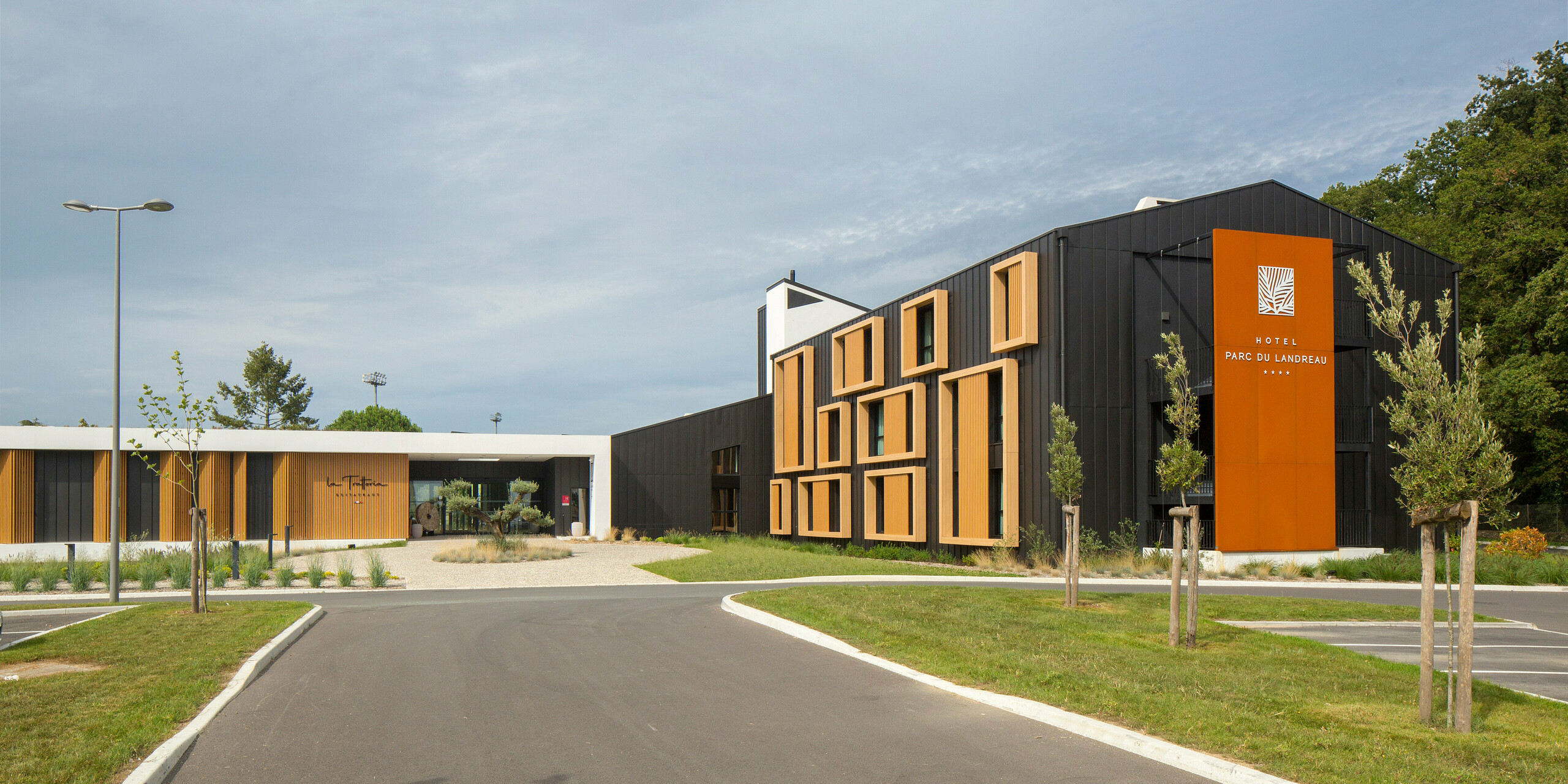 Accès depuis le parking de l'Hôtel Parc du Landreau aux Herbiers, France. Le bâtiment moderne est revêtu du système de toiture et de façade PREFALZ en gris noir. L'architecture moderne est complétée par des cadres en bois marquants autour des fenêtres, qui se fondent harmonieusement dans le revêtement robuste en aluminium. Le vaste parvis, les espaces verts bien entretenus et le design intemporel soulignent l'atmosphère accueillante de l'hôtel.