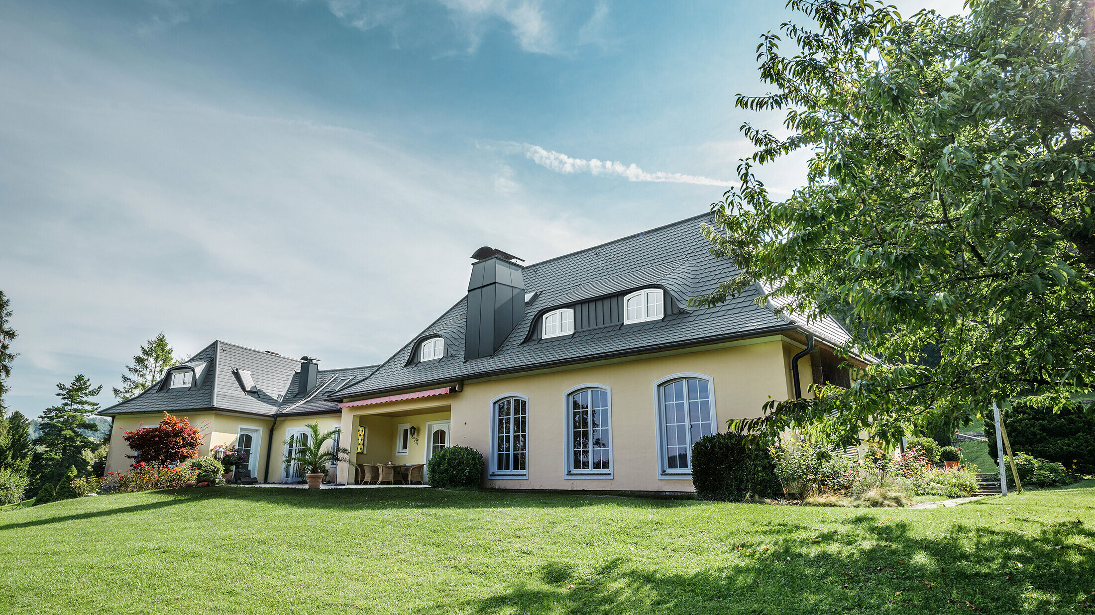 Maison individuelle classique avec lucarne arrondie, recouverte des bardeaux de toiture PREFA résistants aux tempêtes, avec garantie matériau de 40 ans.