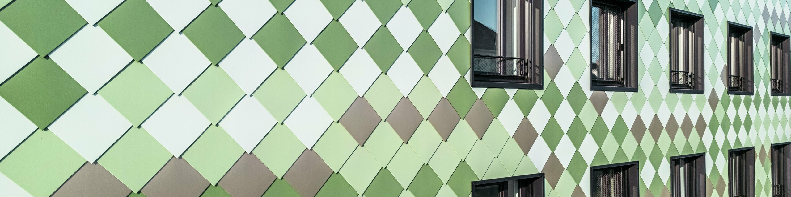 Revêtement de façade moderne PREFA avec losanges PREFA dans des couleurs spéciales spécialement développées : vert olive, vert gris, vert réséda, vert opale, ivoire clair, blanc perle et bronze clair. Le bâtiment résidentiel et commercial « Pick-Nick » à Dresde impressionne par son esthétique unique, sa résistance aux intempéries, sa durabilité et sa qualité sans entretien. La conception stylistique et la variété des couleurs mettent des accents architecturaux et assurent une protection durable.