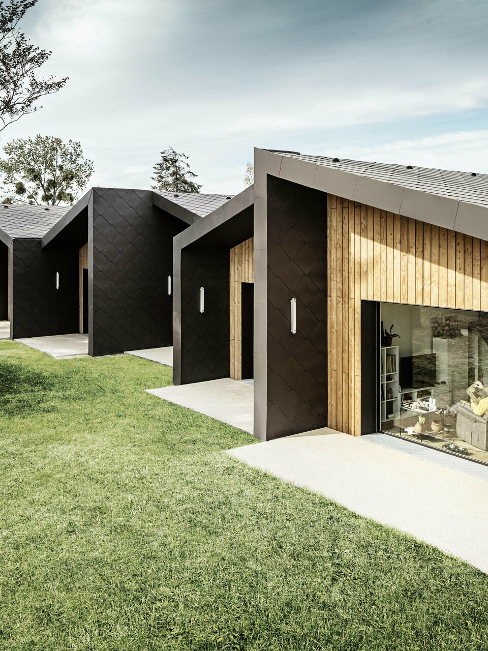 La photo montre une série de bâtiments modernes et minimalistes avec une façade en losanges de toiture et de façade PREFA 44 × 44 de couleur brun noisette. La structure en losange du revêtement s'harmonise avec les éléments en bois chaleureux qui encadrent l'entrée et les grandes façades vitrées. Les bâtiments sont intégrés dans un paysage verdoyant et bien entretenu, donnant une impression calme et accueillante. Le design allie fonctionnalité et architecture esthétique.