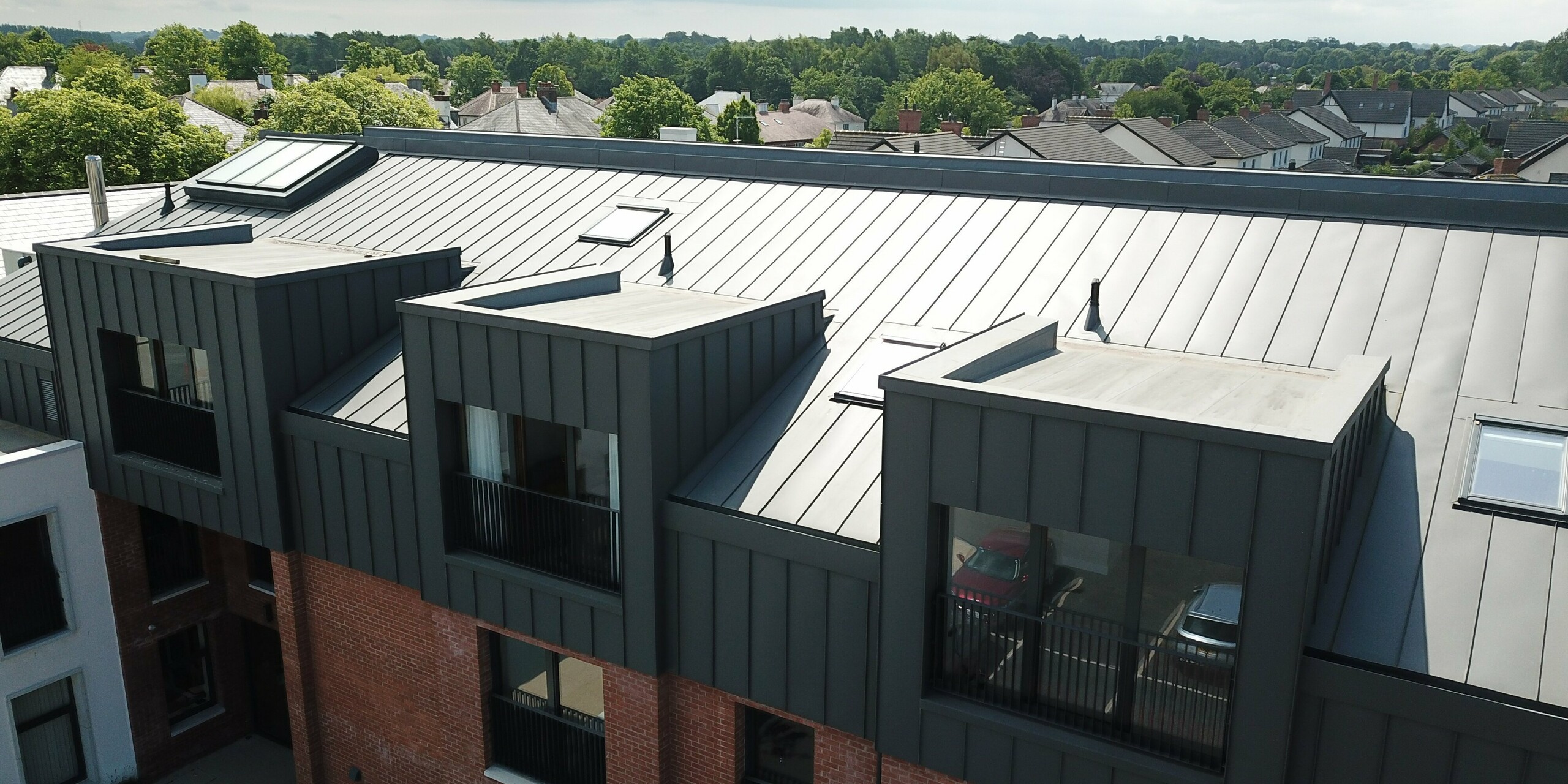 Vue aérienne du toit de Harberton Hall à Belfast, en Irlande du Nord. Le bâtiment moderne destiné aux résidences pour personnes âgées associe l'aluminium PREFALZ en P.10 gris sombre à une façade en brique classique. Les lucarnes à toit plat avec leurs grandes fenêtres assurent des intérieurs lumineux. Le bardage à joint debout crée une combinaison harmonieuse de design contemporain et de construction traditionnelle.