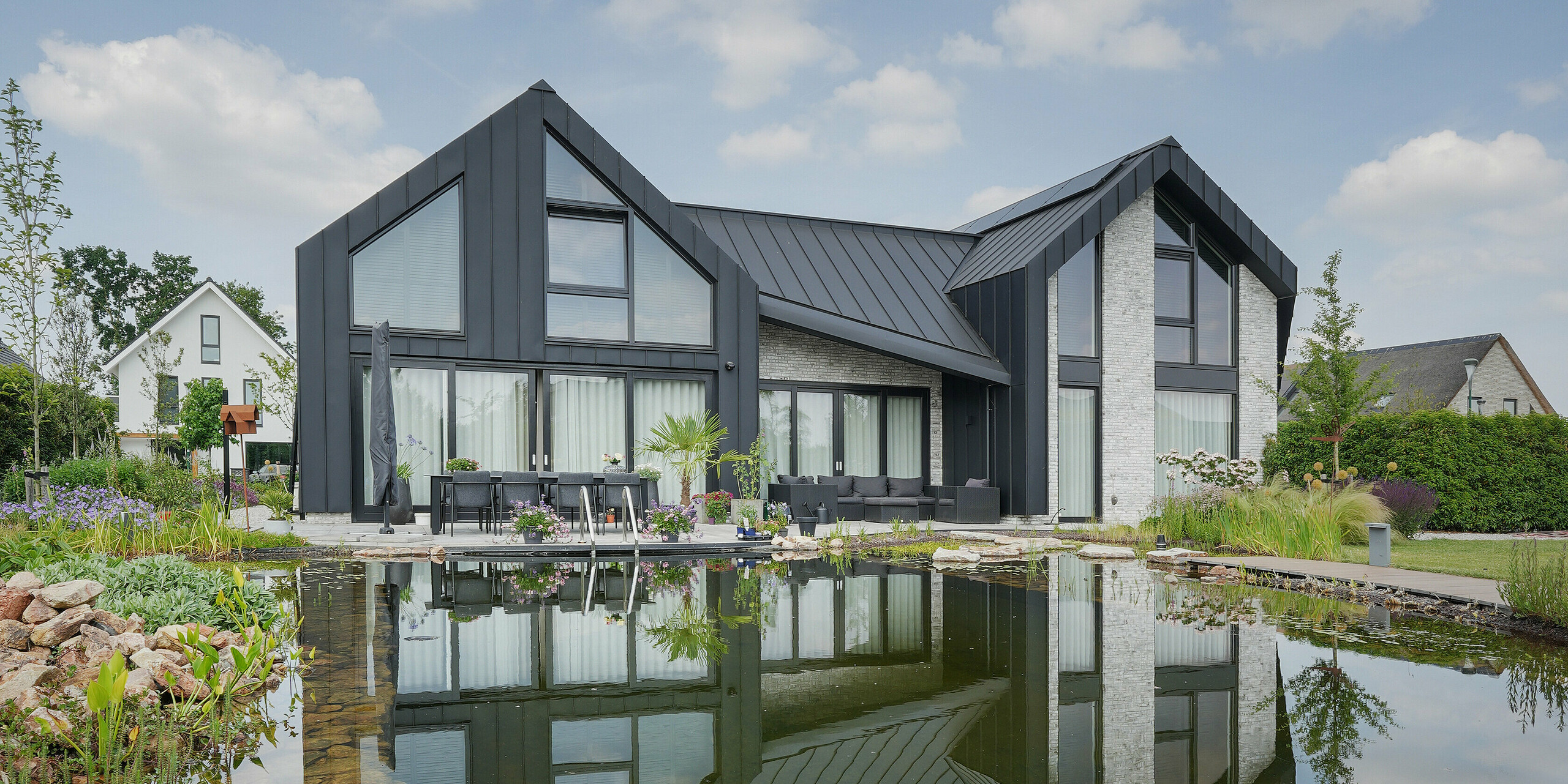 Image idyllique d'une maison familiale à Veenendaal, Pays-Bas, se reflétant dans un étang de jardin aux eaux claires. Le système de toiture et de façade sombre de PREFALZ contraste avec le ciel bleu clair, tandis que l'étang reflète la maison et la nature environnante, soulignant l'atmosphère paisible. Un jardin bien entretenu avec une multitude de plantes et de sièges invite à la détente et complète l'image d'une esthétique d'habitation harmonieuse en lien avec la nature.