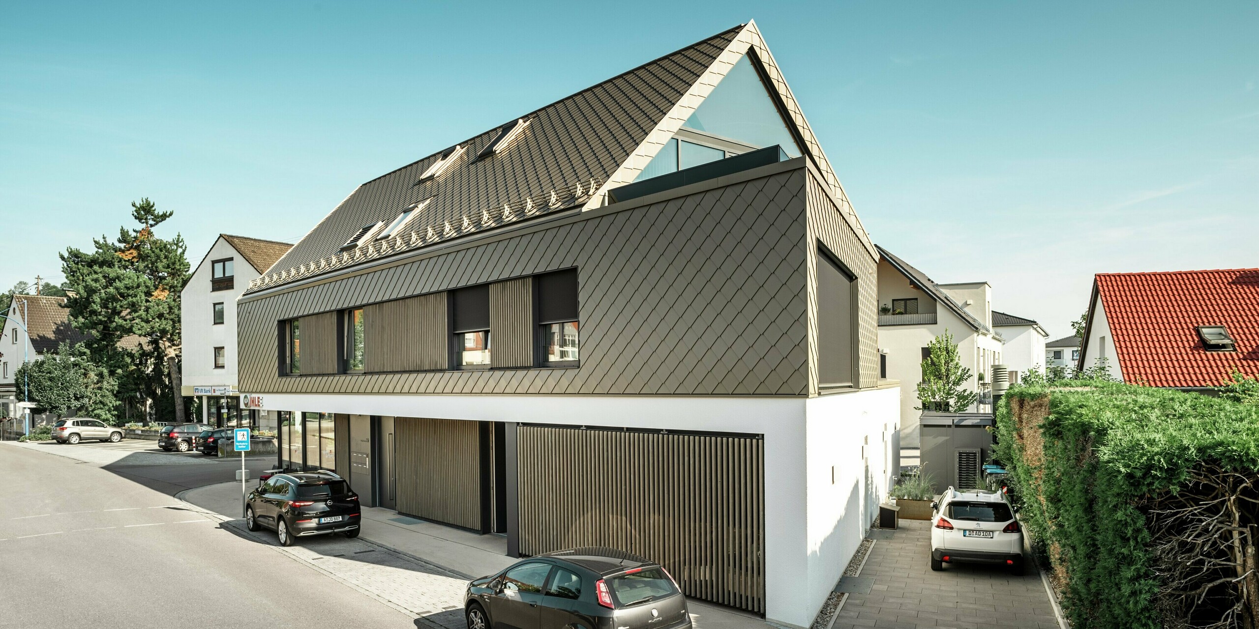 Arrière d'un immeuble d'appartements moderne à Leitershofen, en Allemagne, avec losanges de toiture et de façade 29x29 PREFA en bronze. Le bâtiment combine une façade en crépi blanc au rez-de-chaussée avec des éléments en bois élégants et de grandes surfaces vitrées. La façade de l'étage supérieur et le toit ont été revêtus de robustes losanges en aluminium. Le revêtement en tôle de haute qualité garantit une surface durable et résistante aux intempéries et un design intemporel.