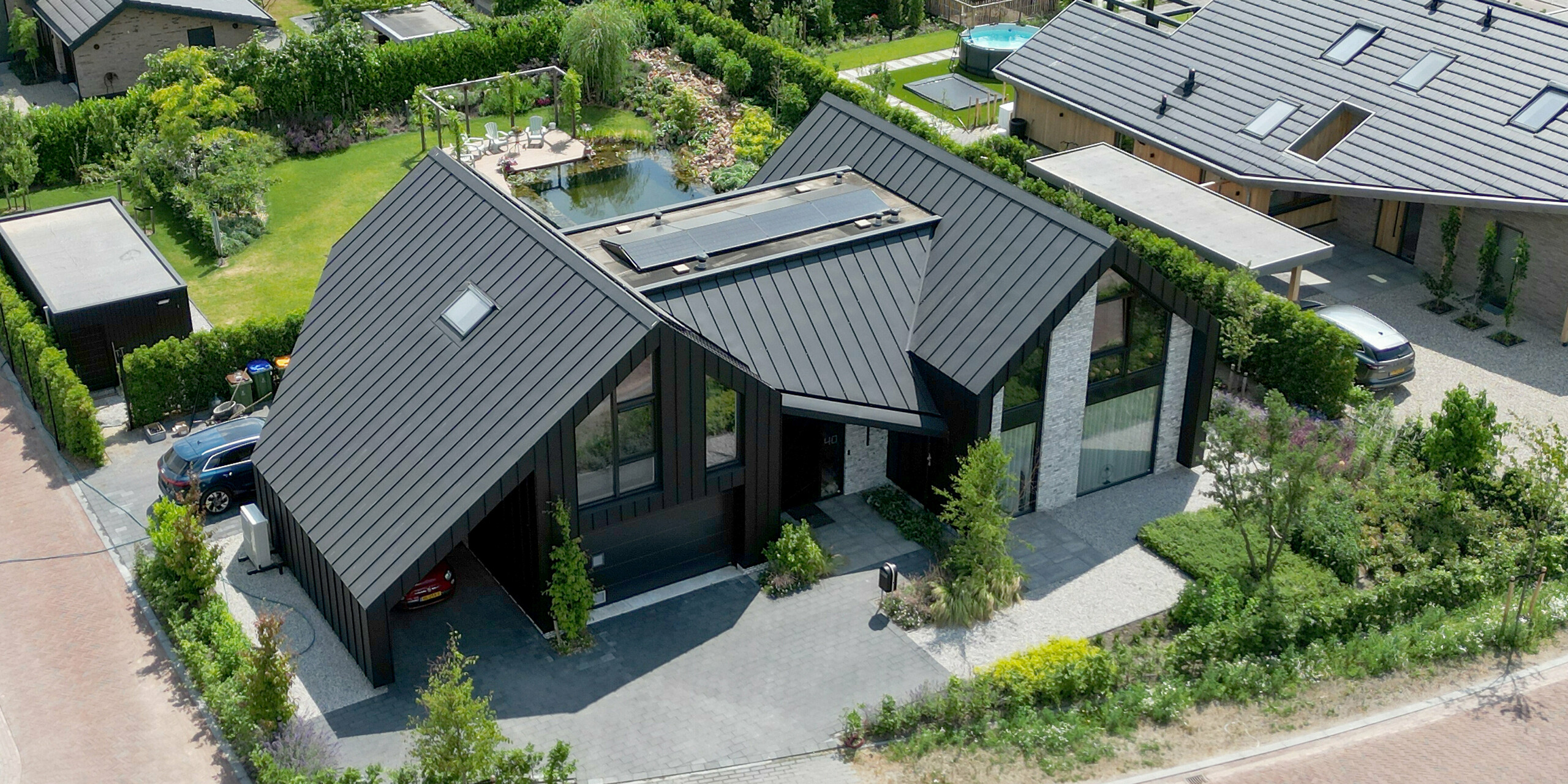 Vue aérienne d'une impressionnante maison individuelle à Veenendaal, aux Pays-Bas, qui se distingue par son toit noir PREFALZ et s'intègre harmonieusement dans le paysage de jardin environnant. La maison est dotée d'un toit à deux pans moderne et constitue un exemple parfait d'architecture moderne grâce à ses lignes épurées et à ses grandes surfaces vitrées encadrées de verdure vivante. L'aménagement paysager bien pensé, avec une allée et un jardin privés, souligne l'esthétique bien pensée de la maison.