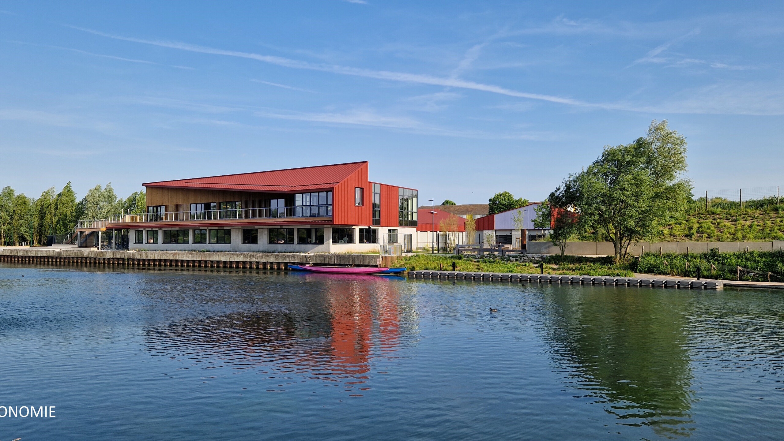 Vue éloignée du stade nautique d'Arras, la teinte rouge oxyde de l'aluminium Prefa se reflète dans la rivière