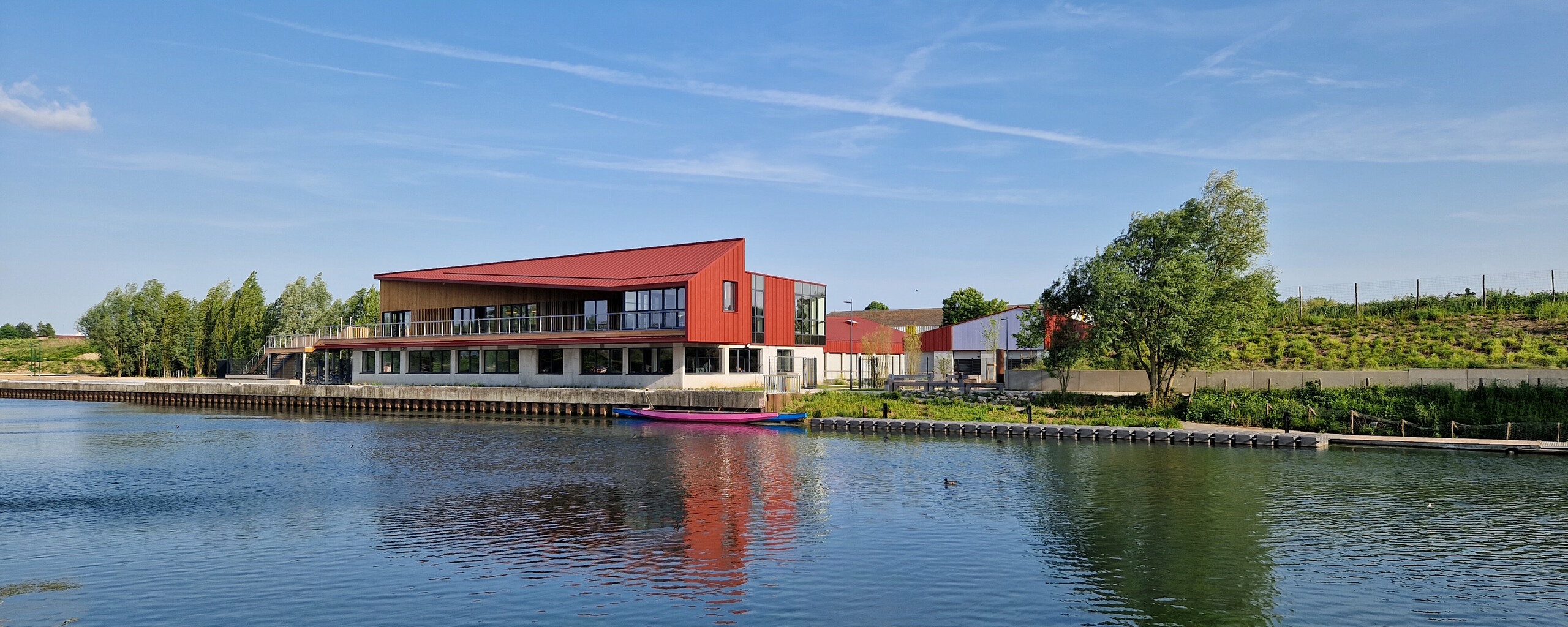 Vue éloignée du stade nautique d'Arras, la teinte rouge oxyde de l'aluminium Prefa se reflète dans la rivière