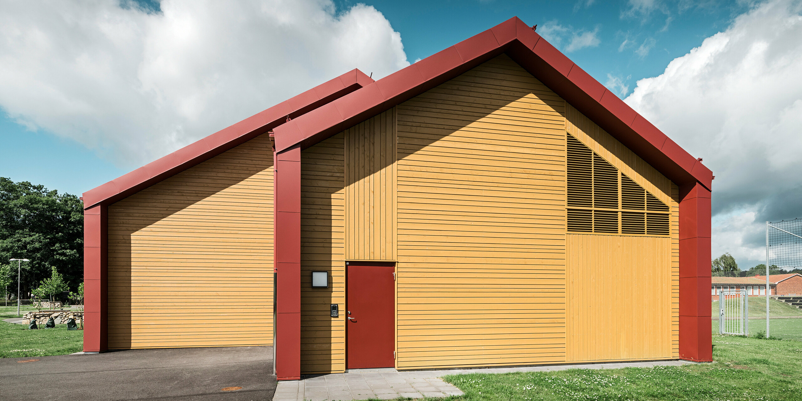 Pignon de la cantine scolaire Restaurang Gröna Hjärtat à Broby, en Suède. La photo montre la façade du bâtiment avec une combinaison d'aluminium PREFALZ rouge oxyde et de revêtement en bois naturel. Les lignes claires de la façade et le contraste de couleurs entre le revêtement en tôle rouge et les tons chauds du bois soulignent le design moderne du bâtiment. Le pignon confère au bâtiment une présence architecturale saisissante et montre l'intégration réussie de matériaux durables et de haute qualité et d'une conception esthétique.